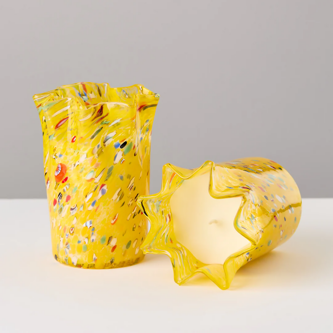 A custom hand-blown yellow candle holder with multicolor speckles, alongside a candle that has been melted to reveal the wax inside, placed on a white surface,  available  to buy on the-salon.com. SALON curates the finest , unique, handmade pieces from the interior industry's best makers and brands. Shop luxury furniture, lighting, rugs, art, antiques and vintage pieces and accessories from SALON interior designed rooms.