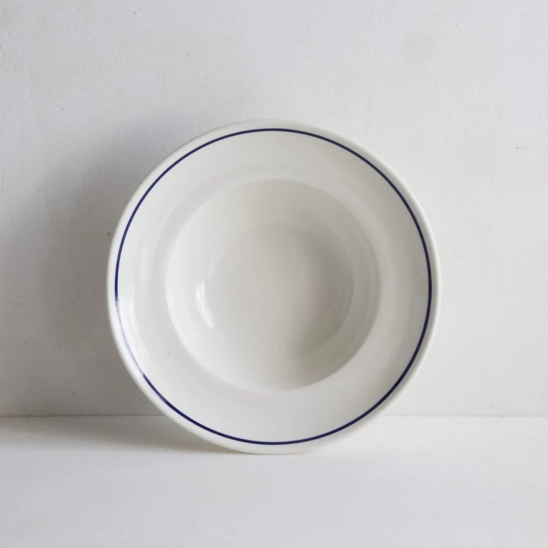 A white porcelain deep bowl with a classic cobalt blue line along the rim, against a plain background, available  to buy on the-salon.com. SALON curates the finest , unique, handmade pieces from the interior industry's best makers and brands. Shop luxury furniture, lighting, rugs, art, antiques and vintage pieces and accessories from SALON interior designed rooms.
