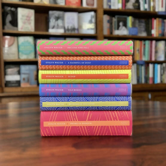 A collection of Evelyn Waugh books republished in hardcover, displayed on a wooden surface with bookshelves in the background,  available  to buy on the-salon.com. SALON curates the finest , unique, handmade pieces from the interior industry's best makers and brands. Shop luxury furniture, lighting, rugs, art, antiques and vintage pieces and accessories from SALON interior designed rooms.