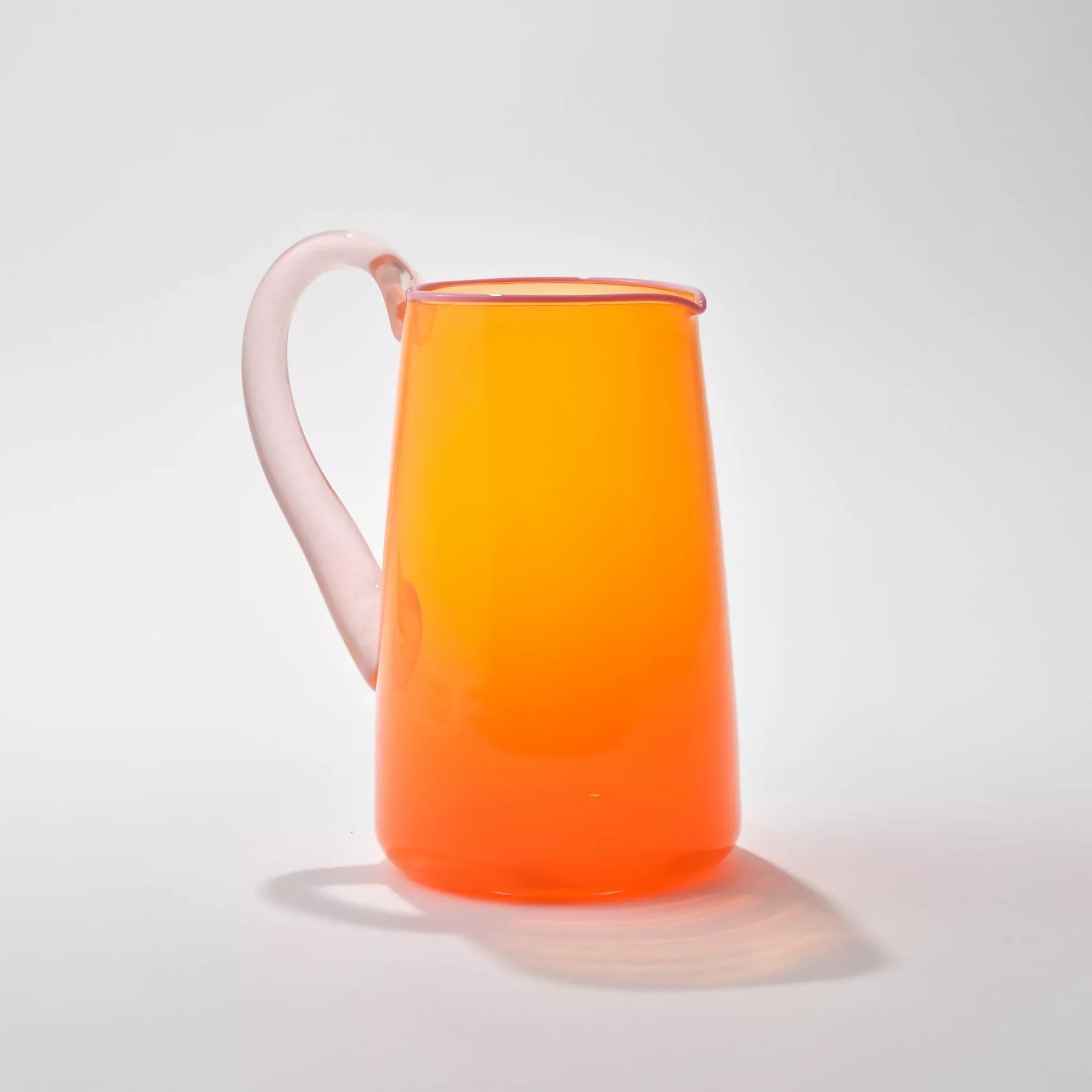 Hand blown, transparent orange glass jug with a blush handle and contrasting pale pink lip on a white background,  available  to buy on the-salon.com. SALON curates the finest , unique, handmade pieces from the interior industry's best makers and brands. Shop luxury furniture, lighting, rugs, art, antiques and vintage pieces and accessories from SALON interior designed rooms.