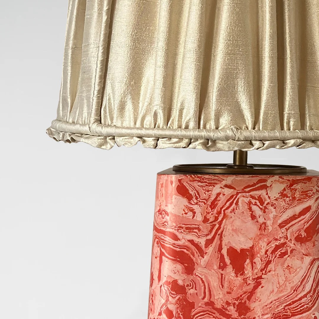 A close up of a red marbled base table lamp with brass detailing, topped with a gold pleated shade,  available  to buy on the-salon.com. SALON curates the finest , unique, handmade pieces from the interior industry's best makers and brands. Shop luxury furniture, lighting, rugs, art, antiques and vintage pieces and accessories from SALON interior designed rooms.