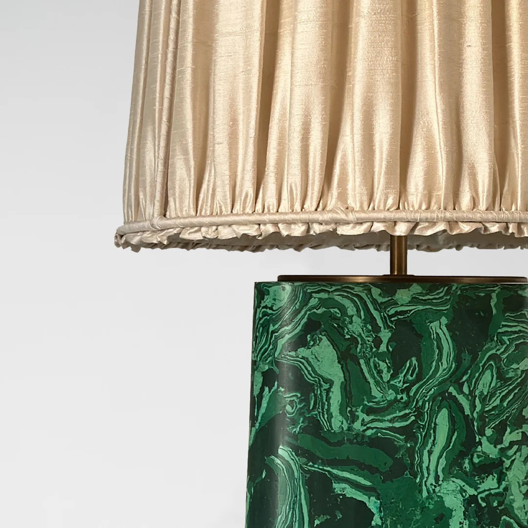 A close up of a  green marbled base table lamp with brass detailing, topped with a gold pleated shade,  available  to buy on the-salon.com. SALON curates the finest , unique, handmade pieces from the interior industry's best makers and brands. Shop luxury furniture, lighting, rugs, art, antiques and vintage pieces and accessories from SALON interior designed rooms.