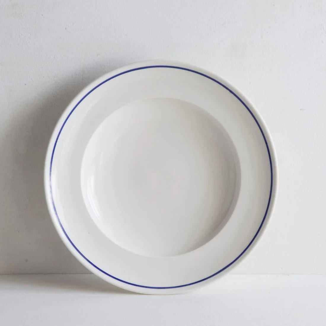 A white porcelain shallow bowl with a cobalt blue line along the rim, against a plain background, available  to buy on the-salon.com. SALON curates the finest , unique, handmade pieces from the interior industry's best makers and brands. Shop luxury furniture, lighting, rugs, art, antiques and vintage pieces and accessories from SALON interior designed rooms.