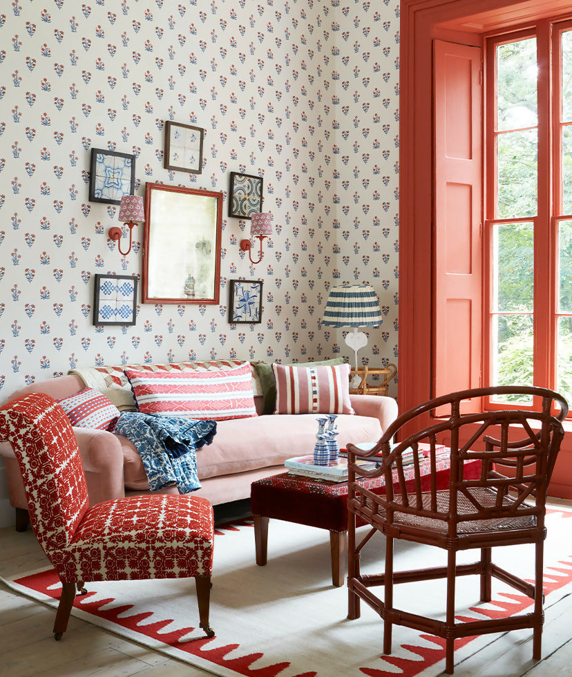Living room with floral wallpaper, colorful furniture, and a large window and two  wall-mounted light fixtures with a red-colored metal arm and a white, patterned lampshade,  available  to buy on the-salon.com. SALON curates the finest , unique, handmade pieces from the interior industry's best makers and brands. Shop luxury furniture, lighting, rugs, art, antiques and vintage pieces and accessories from SALON interior designed rooms.