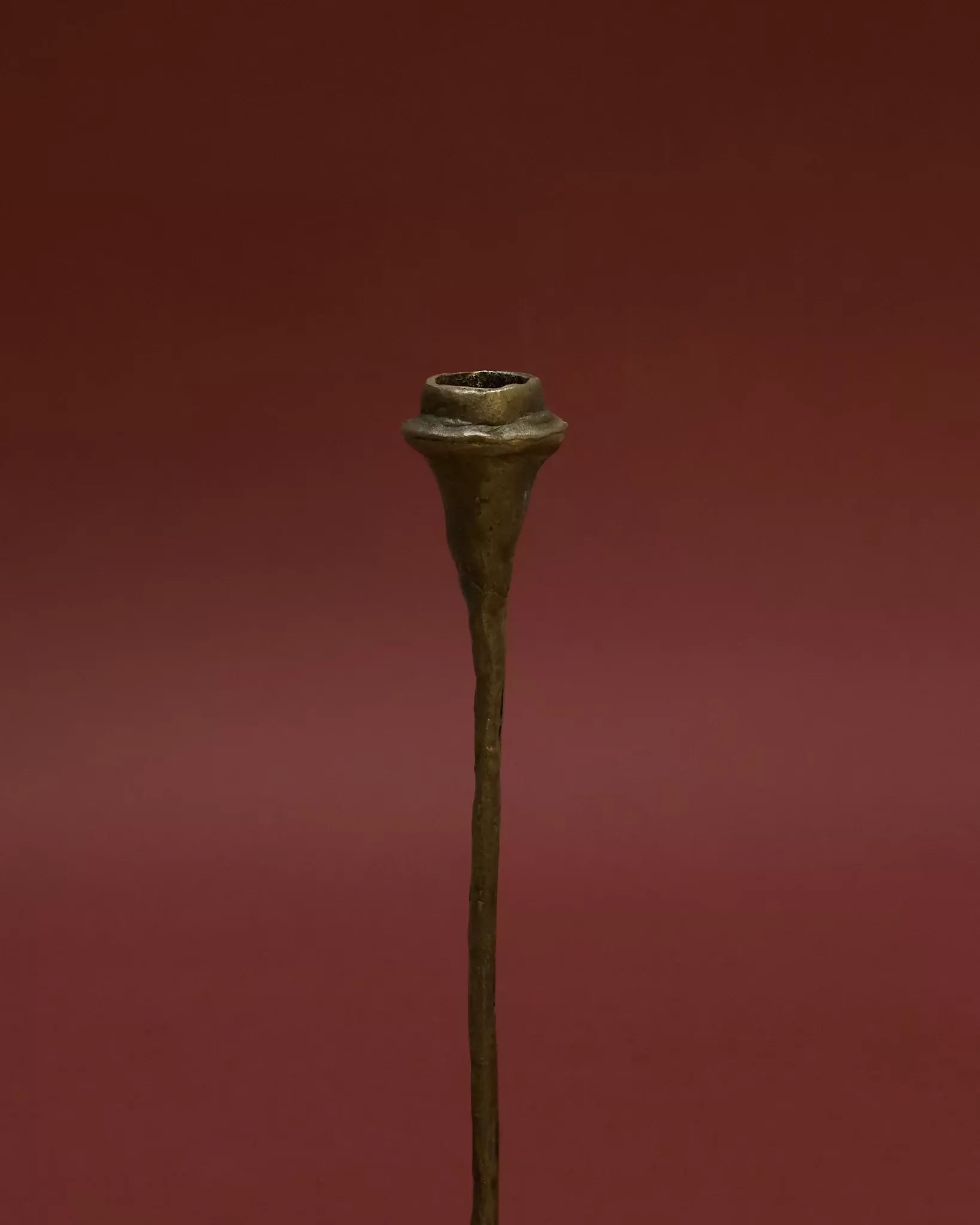 A tall, solid bronze candle holder with a textured finish, standing on a flat surface with a red background,  available  to buy on the-salon.com. SALON curates the finest , unique, handmade pieces from the interior industry's best makers and brands. Shop luxury furniture, lighting, rugs, art, antiques and vintage pieces and accessories from SALON interior designed rooms.