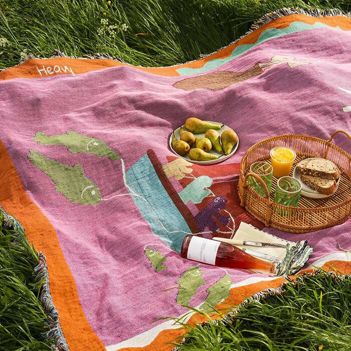 A multicolored throw blanket with a cultural motif laid out on grass, accompanied by a basket of food and drinks,  available  to buy on the-salon.com. SALON curates the finest , unique, handmade pieces from the interior industry's best makers and brands. Shop luxury furniture, lighting, rugs, art, antiques and vintage pieces and accessories from SALON interior designed rooms.