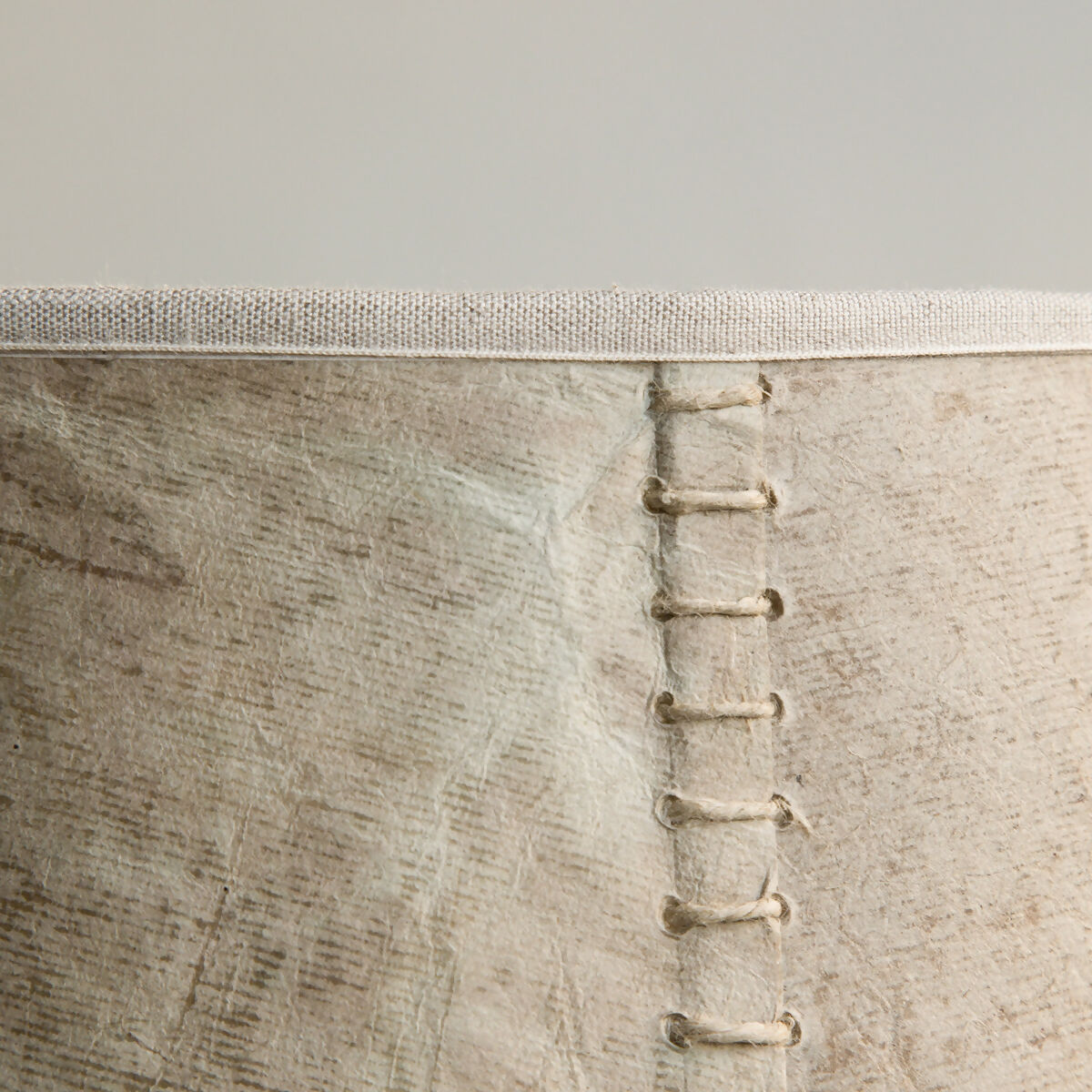 A close up on the stitching on a beige hand stitched tapered lampshade made of Lokta paper with a natural linen trim, against a white background,  available  to buy on the-salon.com. SALON curates the finest , unique, handmade pieces from the interior industry's best makers and brands. Shop luxury furniture, lighting, rugs, art, antiques and vintage pieces and accessories from SALON interior designed rooms.