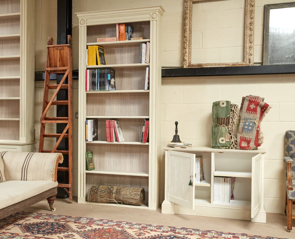 An image of a white bookcase with few books on it in a living room with a white sofa, white walls and frames in the background,  available  to buy on the-salon.com. SALON curates the finest , unique, handmade pieces from the interior industry's best makers and brands. Shop luxury furniture, lighting, rugs, art, antiques and vintage pieces and accessories from SALON interior designed rooms.