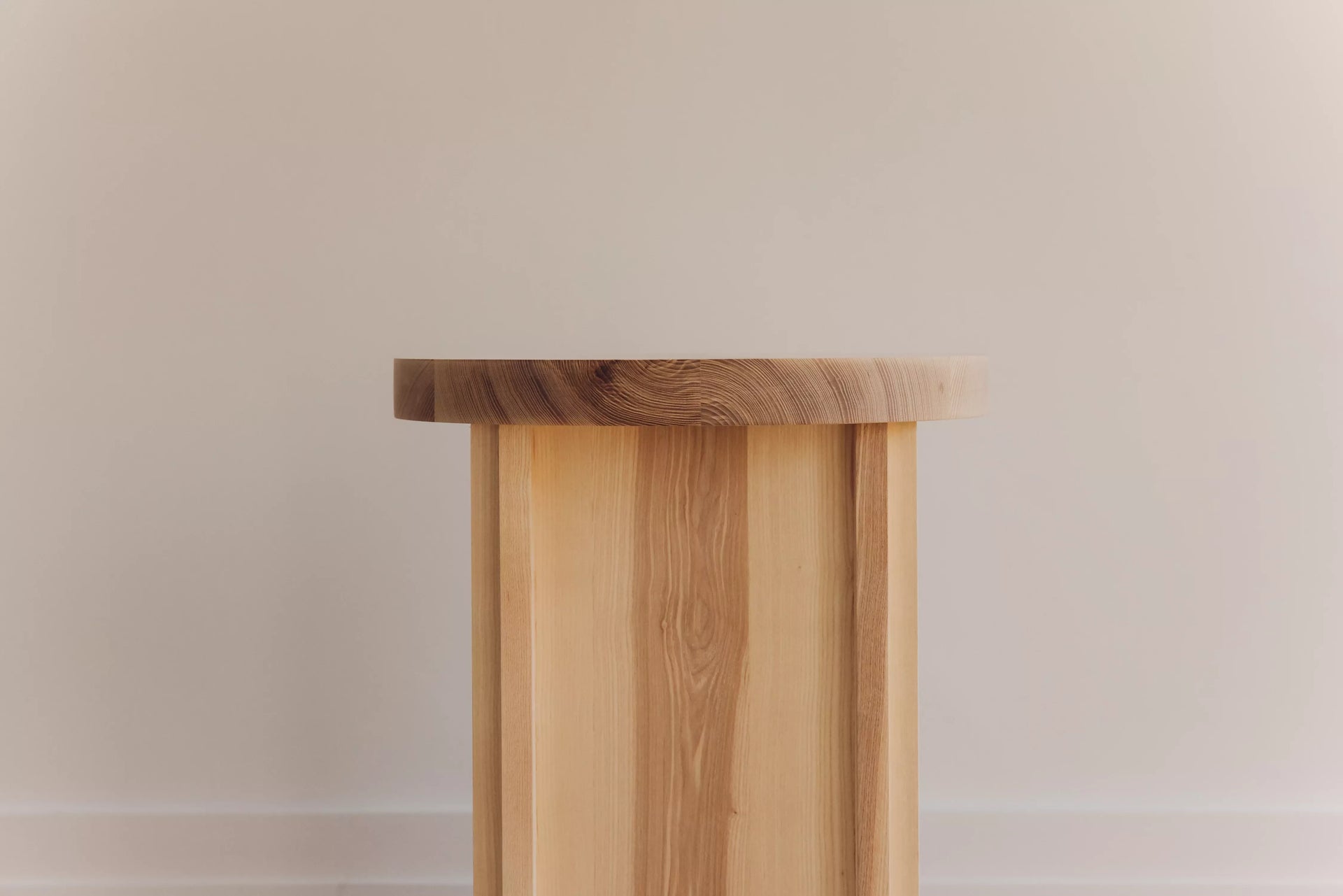 A simple wooden corner side table with a cylindrical shape and visible grain patterns, placed against a neutral background,  available  to buy on the-salon.com. SALON curates the finest , unique, handmade pieces from the interior industry's best makers and brands. Shop luxury furniture, lighting, rugs, art, antiques and vintage pieces and accessories from SALON interior designed rooms.