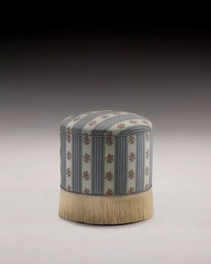 A round footstool upholstered in a multicolored fabric with stripes and floral patterns, featuring a silky champagne fringe at the bottom,  available  to buy on the-salon.com. SALON curates the finest , unique, handmade pieces from the interior industry's best makers and brands. Shop luxury furniture, lighting, rugs, art, antiques and vintage pieces and accessories from SALON interior designed rooms.