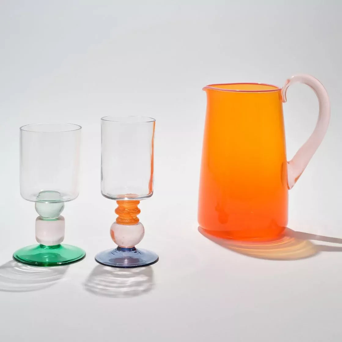 Hand blown, transparent orange glass jug with a blush handle and contrasting pale pink lip on a white background, with two clear glasses with colourful stands,  available  to buy on the-salon.com. SALON curates the finest , unique, handmade pieces from the interior industry's best makers and brands. Shop luxury furniture, lighting, rugs, art, antiques and vintage pieces and accessories from SALON interior designed rooms.