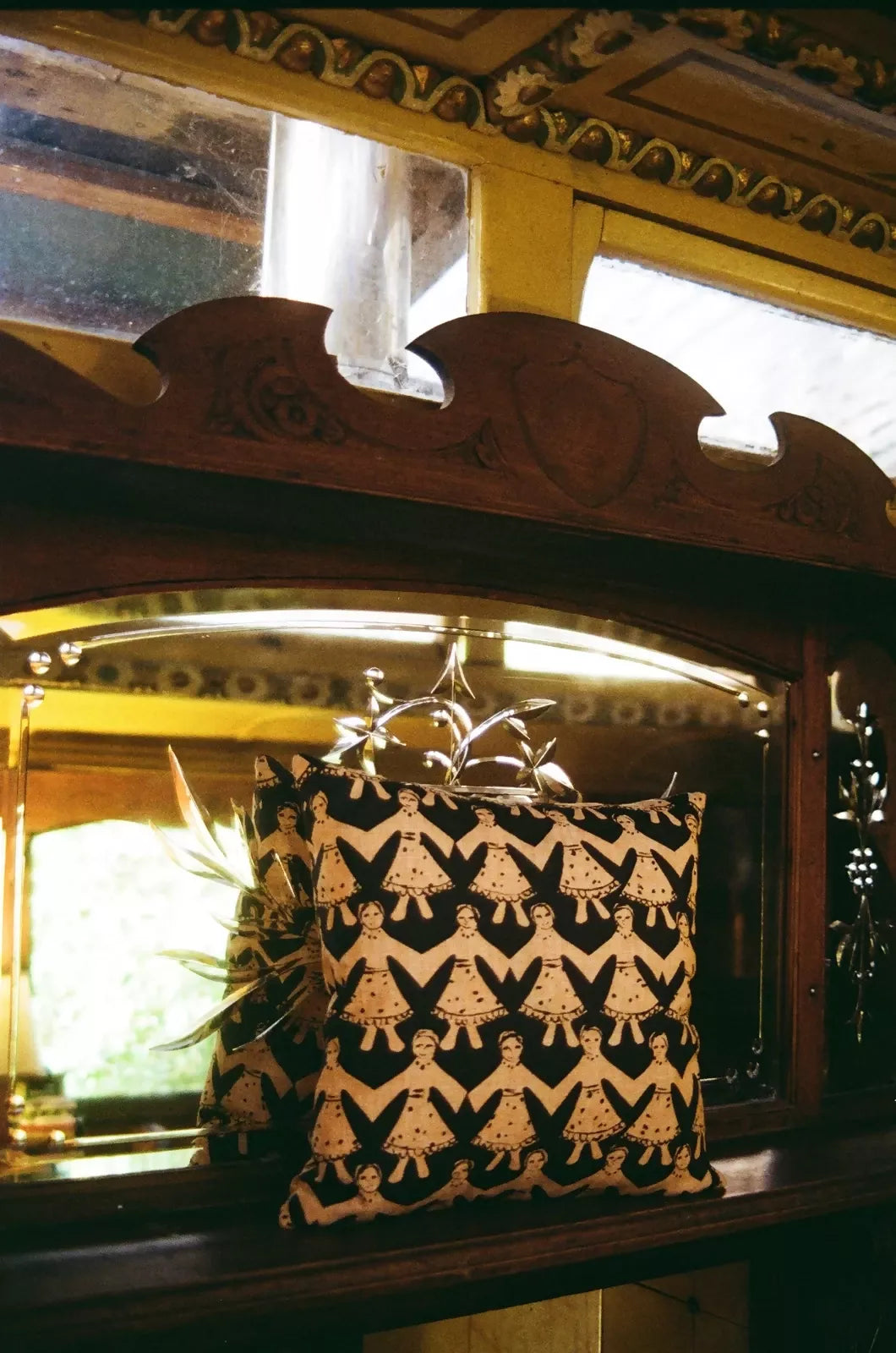 A square-shaped cushion with a geometric pattern, featuring black and gold colors, displayed in front of a window with a ornate wooden frame,  available  to buy on the-salon.com. SALON curates the finest , unique, handmade pieces from the interior industry's best makers and brands. Shop luxury furniture, lighting, rugs, art, antiques and vintage pieces and accessories from SALON interior designed rooms.