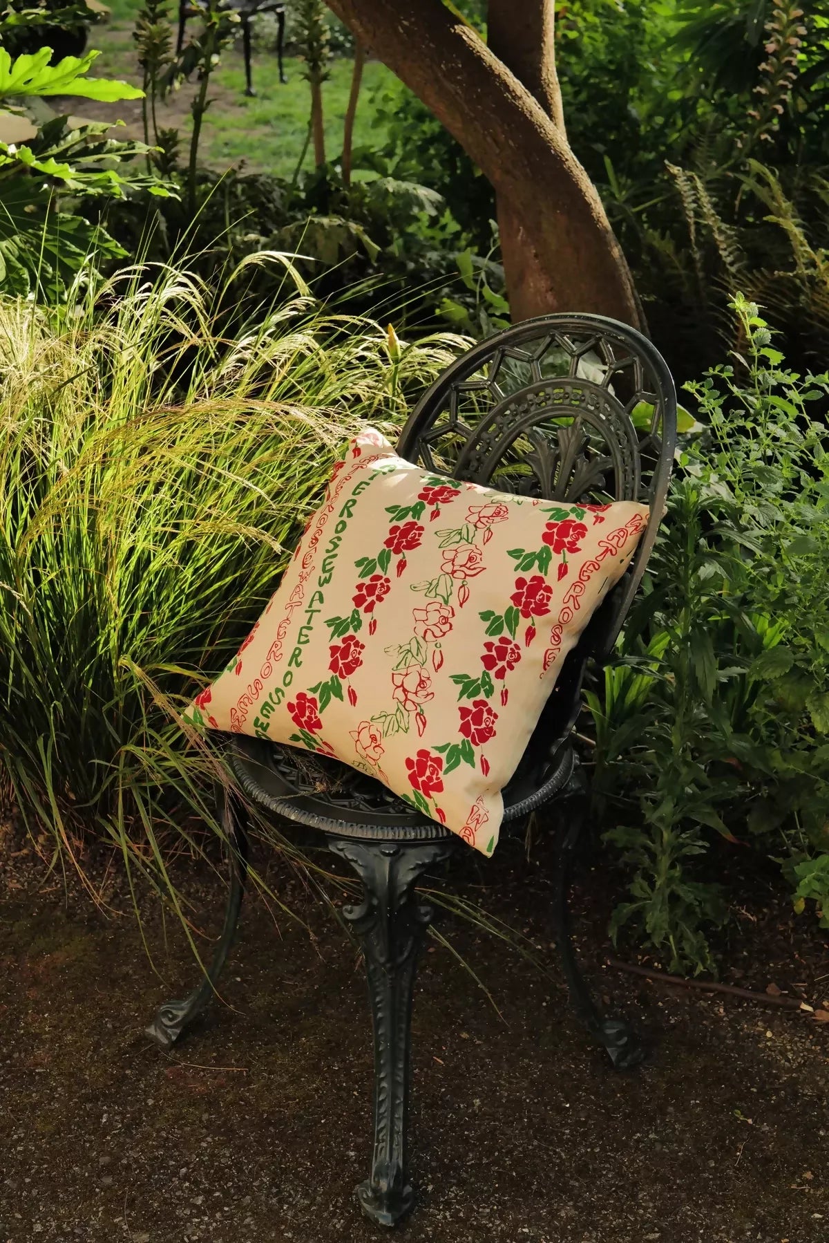 A rectangular cushion with a floral pattern, featuring red flowers and green leaves, resting on a black metal chair,  available  to buy on the-salon.com. SALON curates the finest , unique, handmade pieces from the interior industry's best makers and brands. Shop luxury furniture, lighting, rugs, art, antiques and vintage pieces and accessories from SALON interior designed rooms.