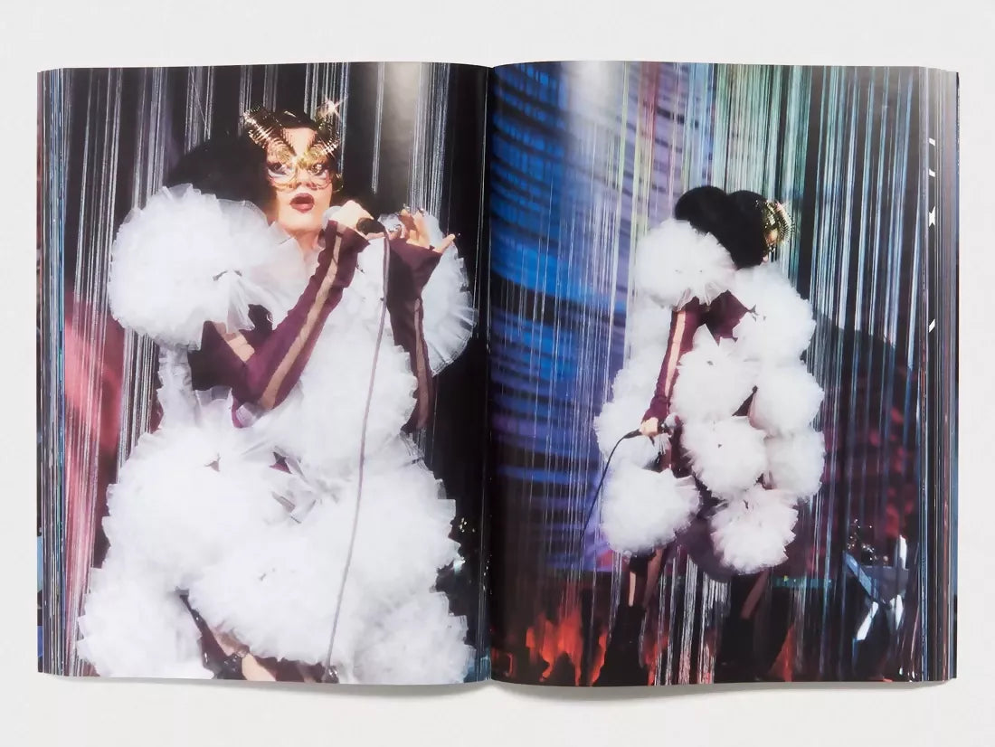 An image of an open book with an image of a woman in the mirror,  looking away wearing a white frilly dress with maroon under, wearing a gold mask with the lights on her, available  to buy on the-salon.com. SALON curates the finest , unique, handmade pieces from the interior industry's best makers and brands. Shop luxury furniture, lighting, rugs, art, antiques and vintage pieces and accessories from SALON interior designed rooms.