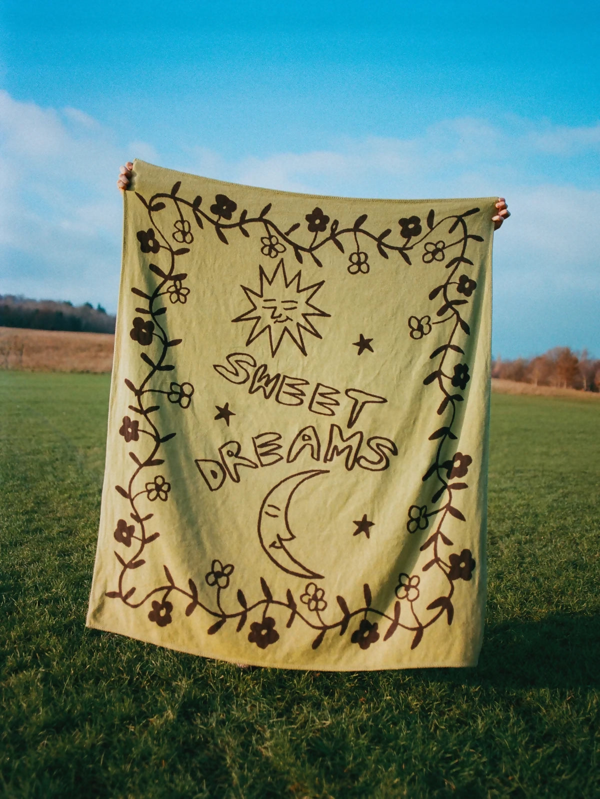 A beige cotton blanket with 'SWEET DREAMS' embroidered on it, displayed outdoors,  available  to buy on the-salon.com. SALON curates the finest , unique, handmade pieces from the interior industry's best makers and brands. Shop luxury furniture, lighting, rugs, art, antiques and vintage pieces and accessories from SALON interior designed rooms.