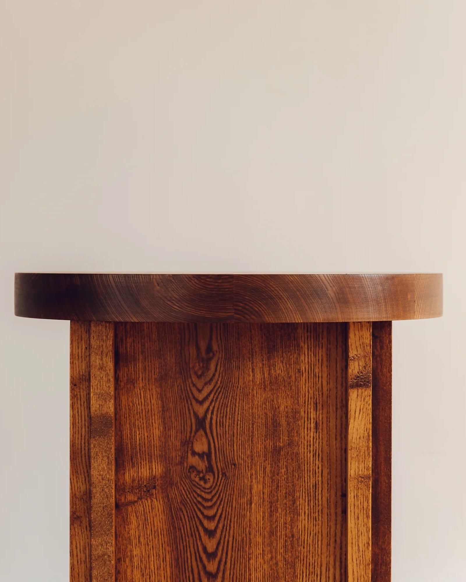 A corner side table made of wood with a visible grain pattern, featuring a cylindrical base and a flat, circular tabletop,  available  to buy on the-salon.com. SALON curates the finest , unique, handmade pieces from the interior industry's best makers and brands. Shop luxury furniture, lighting, rugs, art, antiques and vintage pieces and accessories from SALON interior designed rooms.