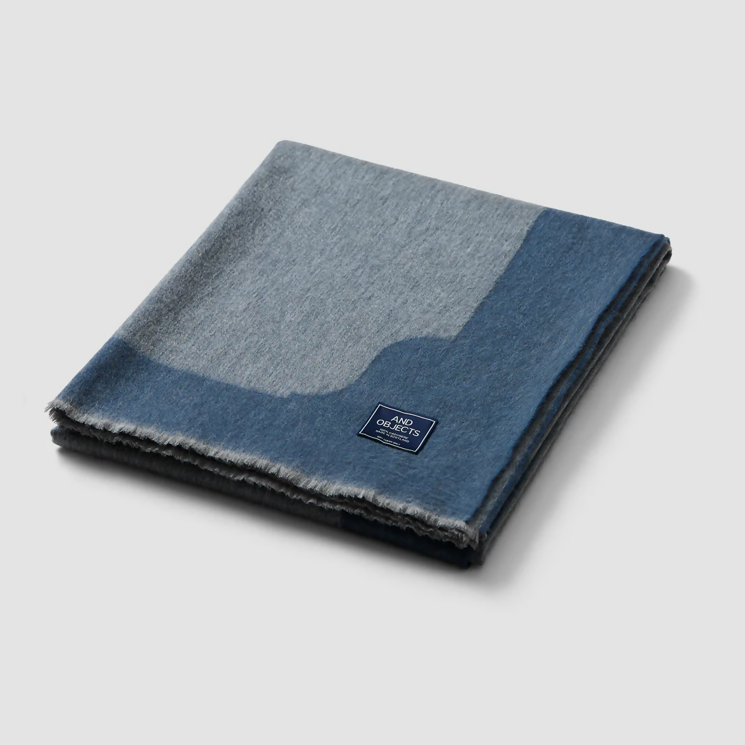 A folded slate blue cashmere throw blanket with a decorative border detail,  available  to buy on the-salon.com. SALON curates the finest , unique, handmade pieces from the interior industry's best makers and brands. Shop luxury furniture, lighting, rugs, art, antiques and vintage pieces and accessories from SALON interior designed rooms.