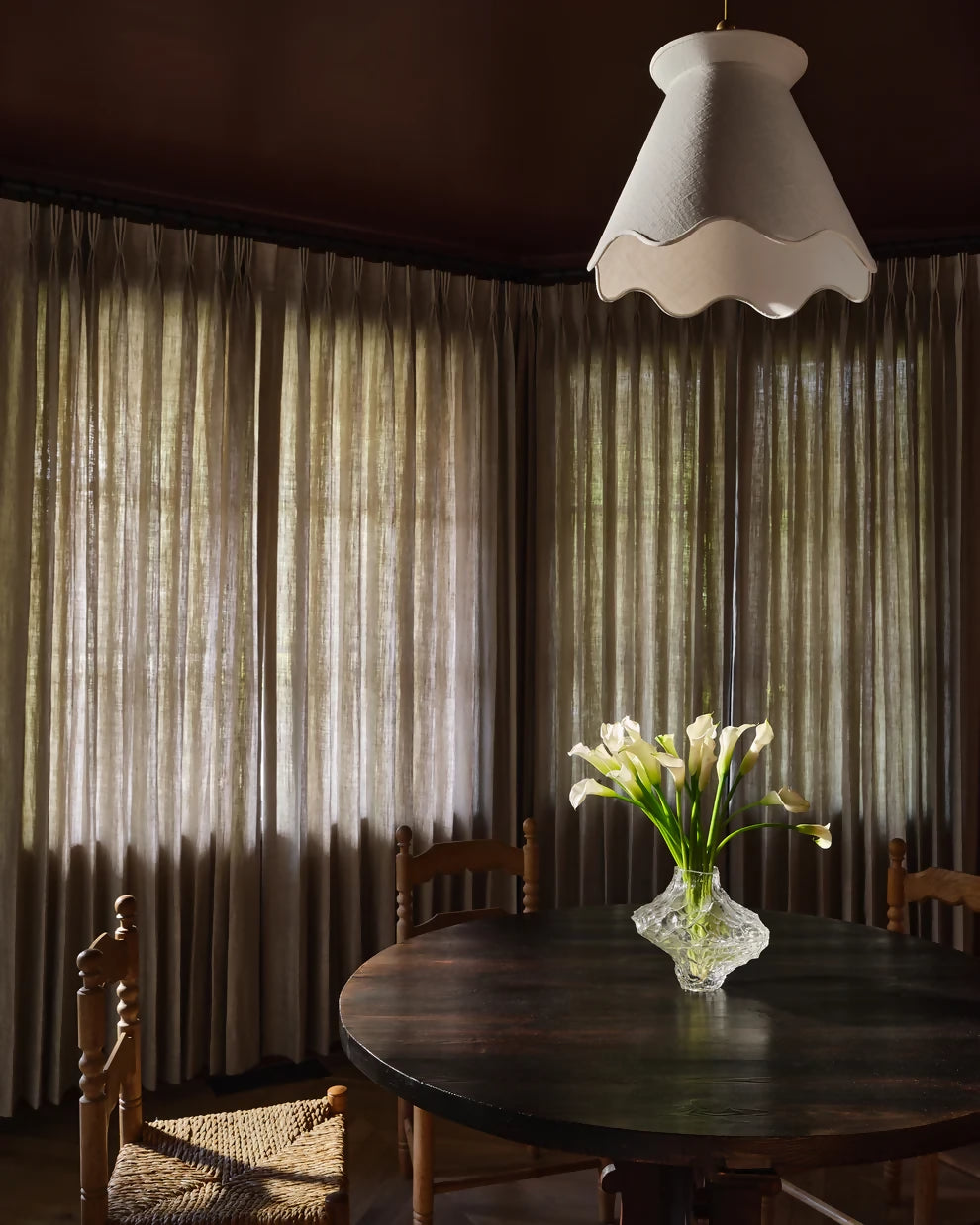 A pendant light with a pleated shade hanging over a dining table with a floral arrangement in the center, with curtains in the background, available  to buy on the-salon.com. SALON curates the finest , unique, handmade pieces from the interior industry's best makers and brands. Shop luxury furniture, lighting, rugs, art, antiques and vintage pieces and accessories from SALON interior designed rooms.