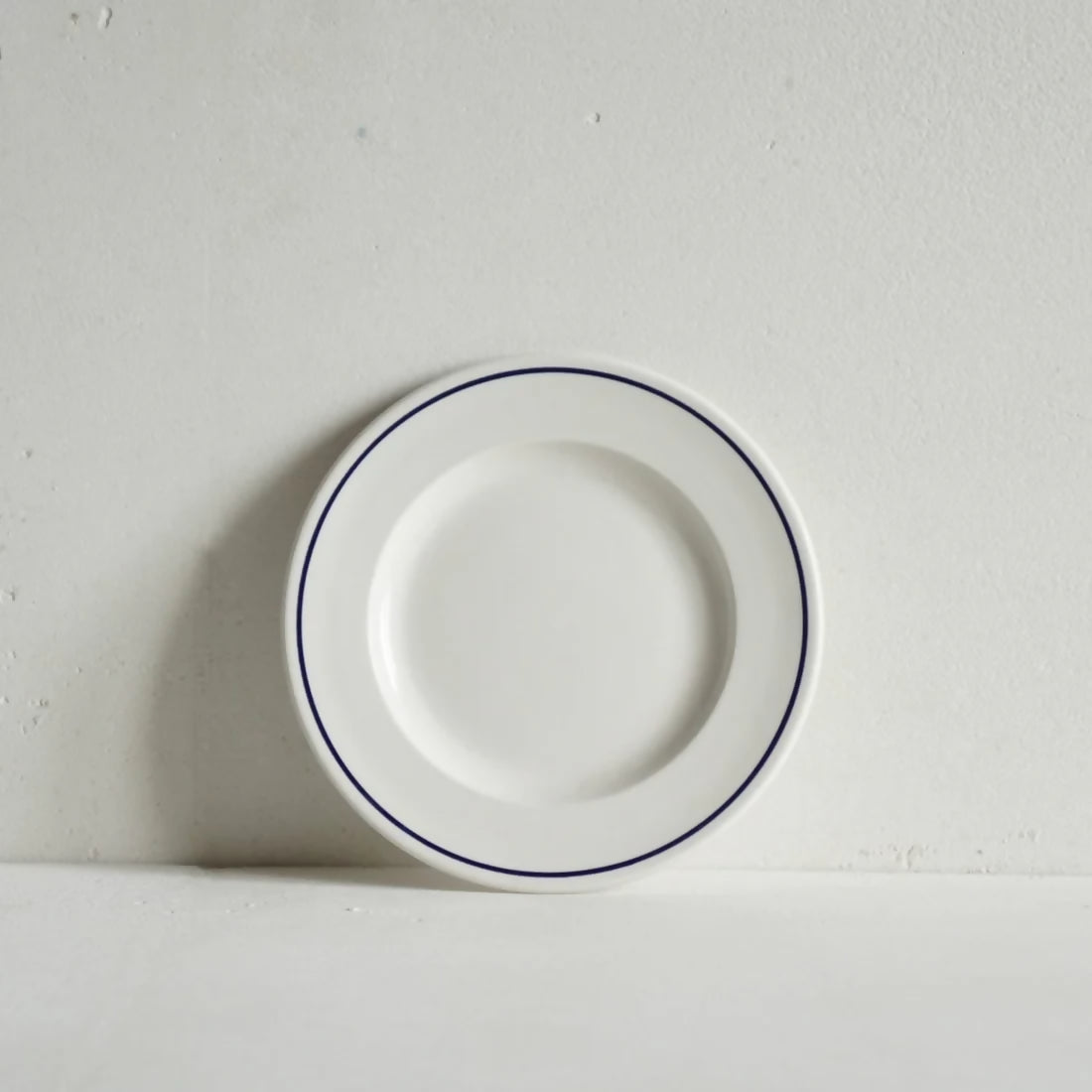 A small round white porcelain side plate with a rich cobalt blue line along the rim, displayed against a white background, available  to buy on the-salon.com. SALON curates the finest , unique, handmade pieces from the interior industry's best makers and brands. Shop luxury furniture, lighting, rugs, art, antiques and vintage pieces and accessories from SALON interior designed rooms.