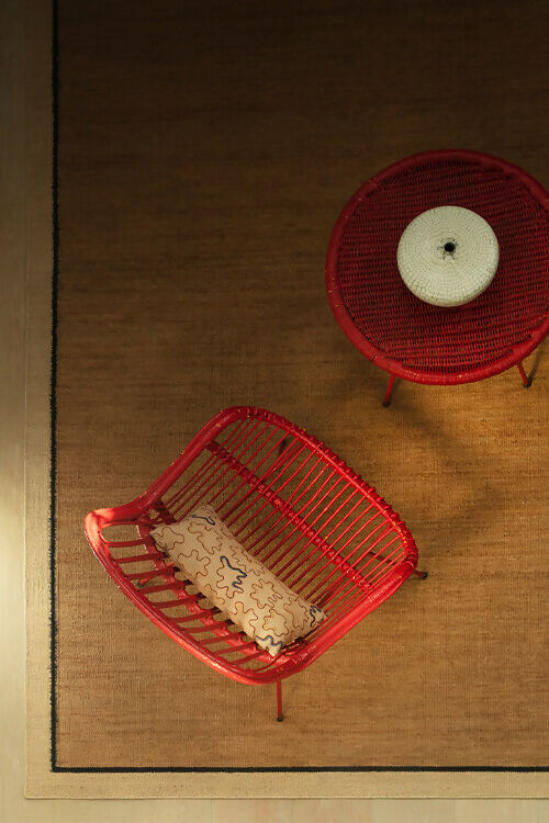 Top view of a flatweave dhurrie rug in a plain beige color with a border, featuring a red circular trivet on top with a white candle, and a red square trivet with a beige object,  available  to buy on the-salon.com. SALON curates the finest , unique, handmade pieces from the interior industry's best makers and brands. Shop luxury furniture, lighting, rugs, art, antiques and vintage pieces and accessories from SALON interior designed rooms.