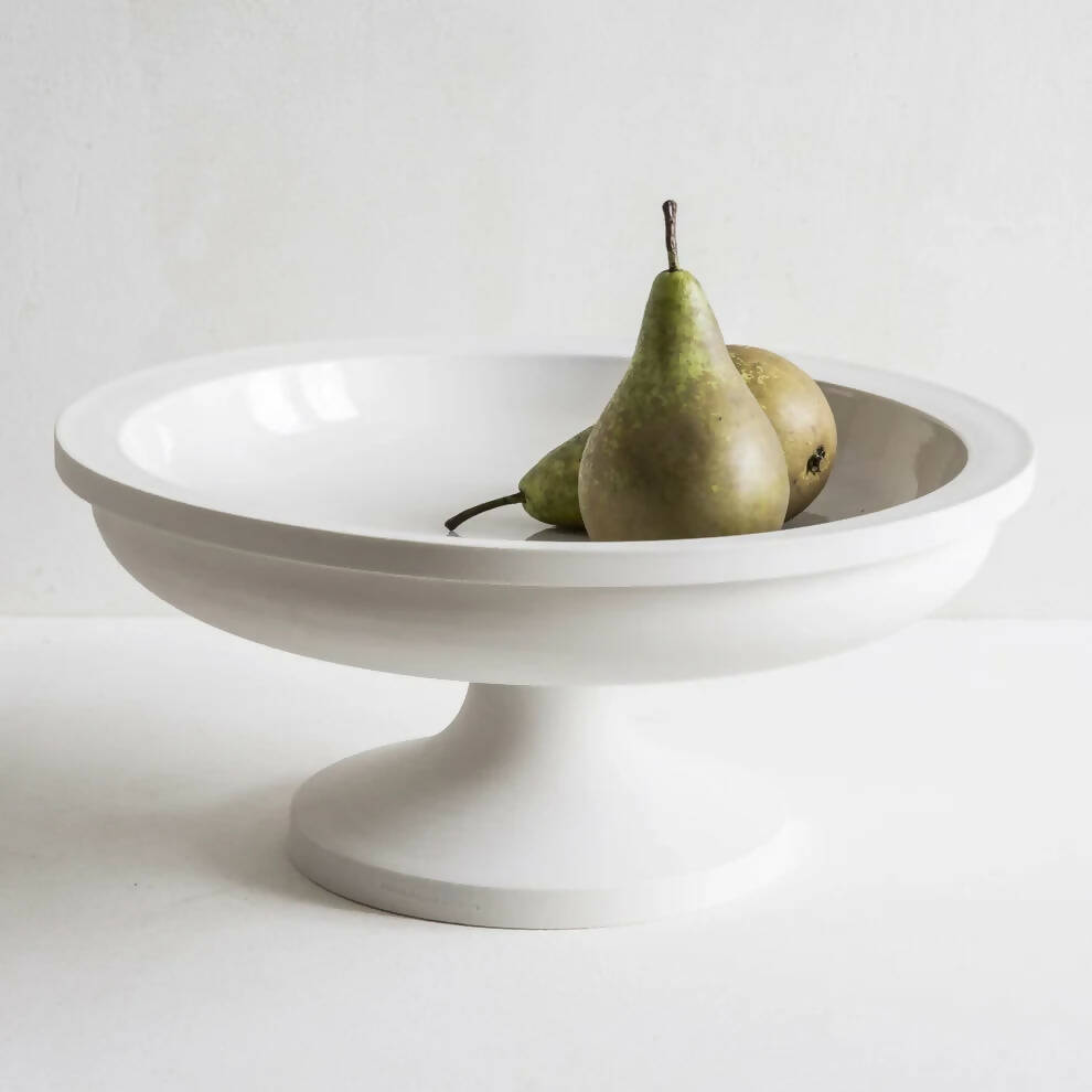 A white porcelain fruit stand with a sculptural foot, containing two pears, on a plain background,  available  to buy on the-salon.com. SALON curates the finest , unique, handmade pieces from the interior industry's best makers and brands. Shop luxury furniture, lighting, rugs, art, antiques and vintage pieces and accessories from SALON interior designed rooms.