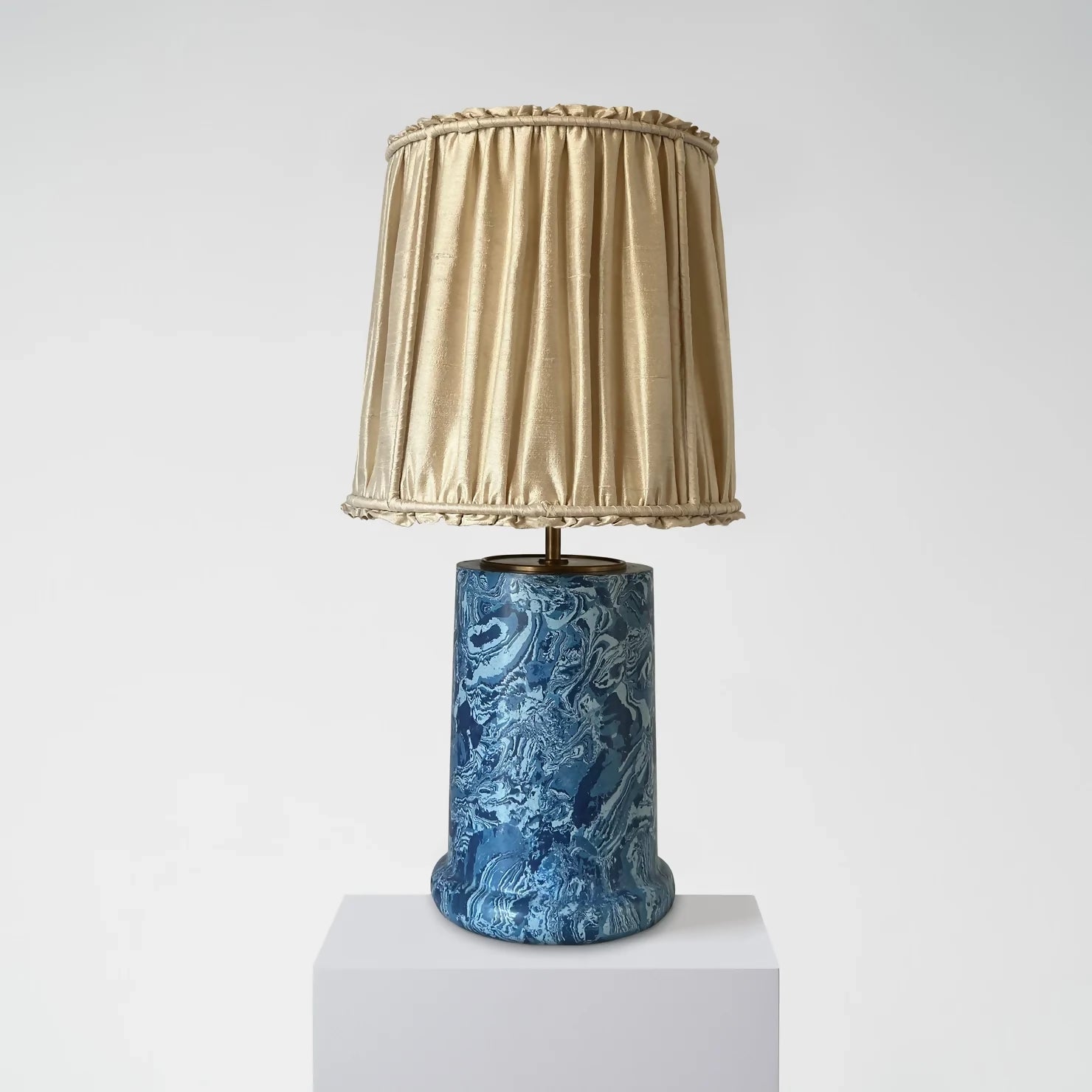 A blue marbled base table lamp with brass detailing, topped with a gold pleated shade,  available  to buy on the-salon.com. SALON curates the finest , unique, handmade pieces from the interior industry's best makers and brands. Shop luxury furniture, lighting, rugs, art, antiques and vintage pieces and accessories from SALON interior designed rooms.