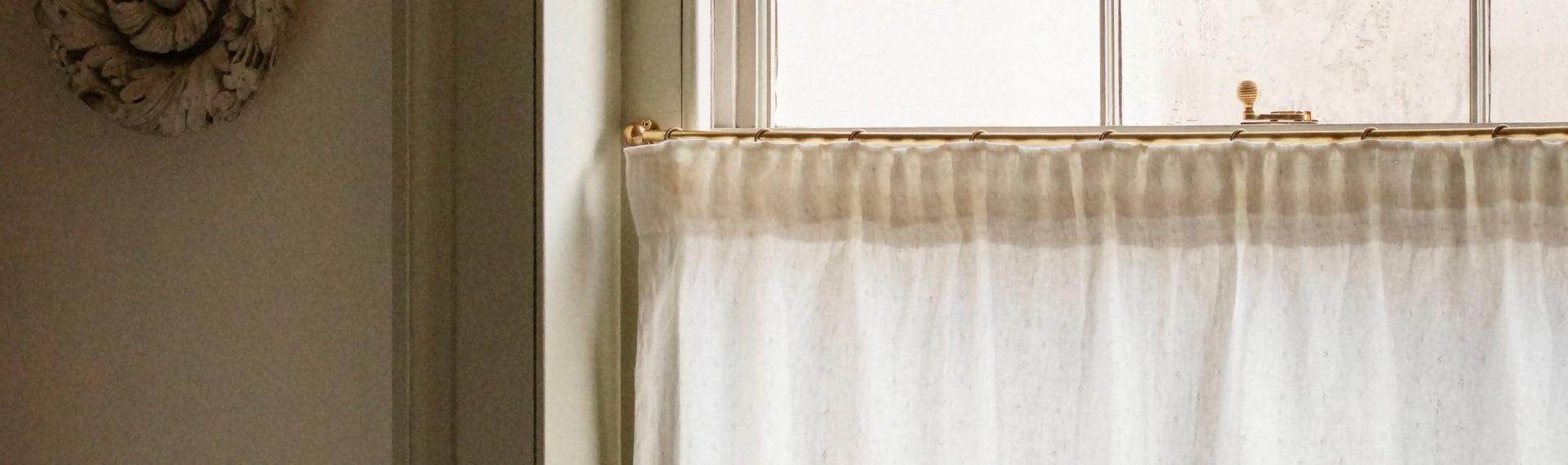 A white cafe curtain made of linen, hanging on a rod, with a window behind it allowing natural light to come through, available  to buy on the-salon.com. SALON curates the finest , unique, handmade pieces from the interior industry's best makers and brands. Shop luxury furniture, lighting, rugs, art, antiques and vintage pieces and accessories from SALON interior designed rooms.