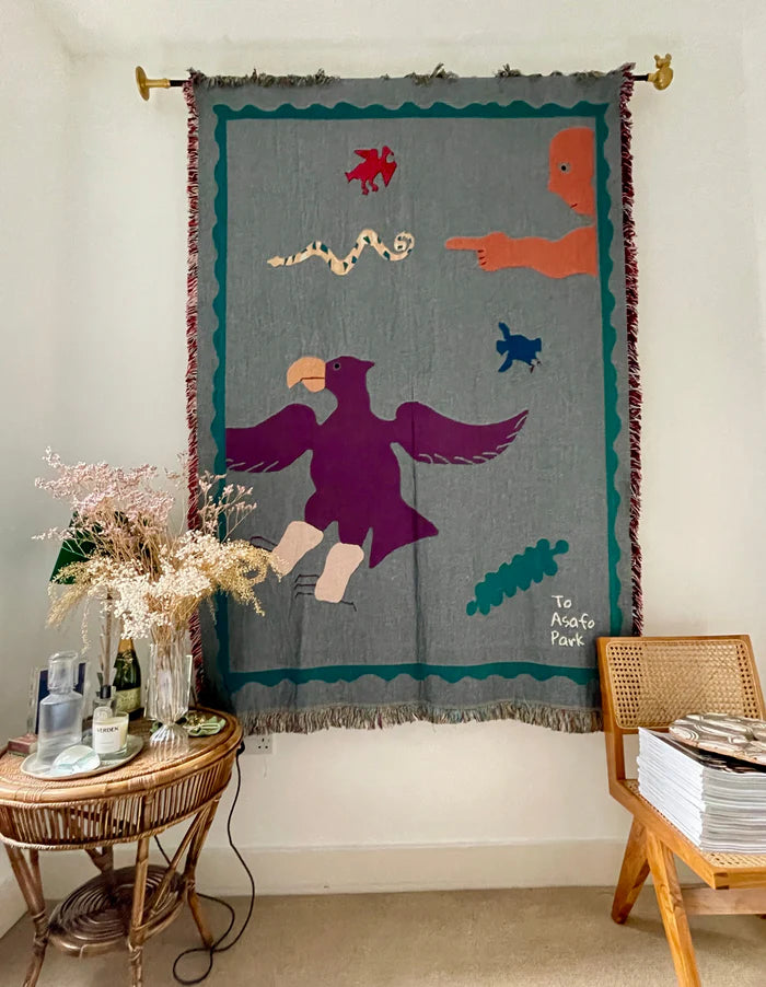A colorful handcrafted blanket with animal and imaginative figures displayed on a wall, titled 'To ASAFO PARK THROW BLANKET BY AMECHI',  available  to buy on the-salon.com. SALON curates the finest , unique, handmade pieces from the interior industry's best makers and brands. Shop luxury furniture, lighting, rugs, art, antiques and vintage pieces and accessories from SALON interior designed rooms.