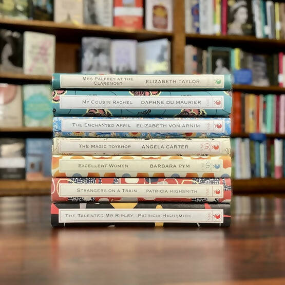 A stack of books from the Virago Modern Classics collection, featuring various cover designs, placed in front of a bookshelf, available  to buy on the-salon.com. SALON curates the finest , unique, handmade pieces from the interior industry's best makers and brands. Shop luxury furniture, lighting, rugs, art, antiques and vintage pieces and accessories from SALON interior designed rooms.
