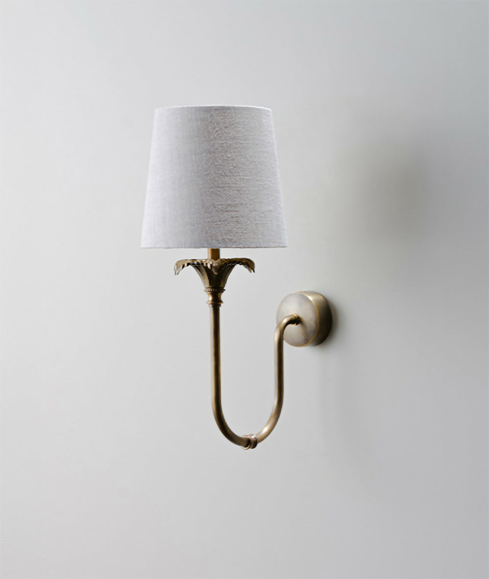 A wall-mounted light fixture with a gold-colored metal arm and a white, solid-patterned lampshade,  available  to buy on the-salon.com. SALON curates the finest , unique, handmade pieces from the interior industry's best makers and brands. Shop luxury furniture, lighting, rugs, art, antiques and vintage pieces and accessories from SALON interior designed rooms.