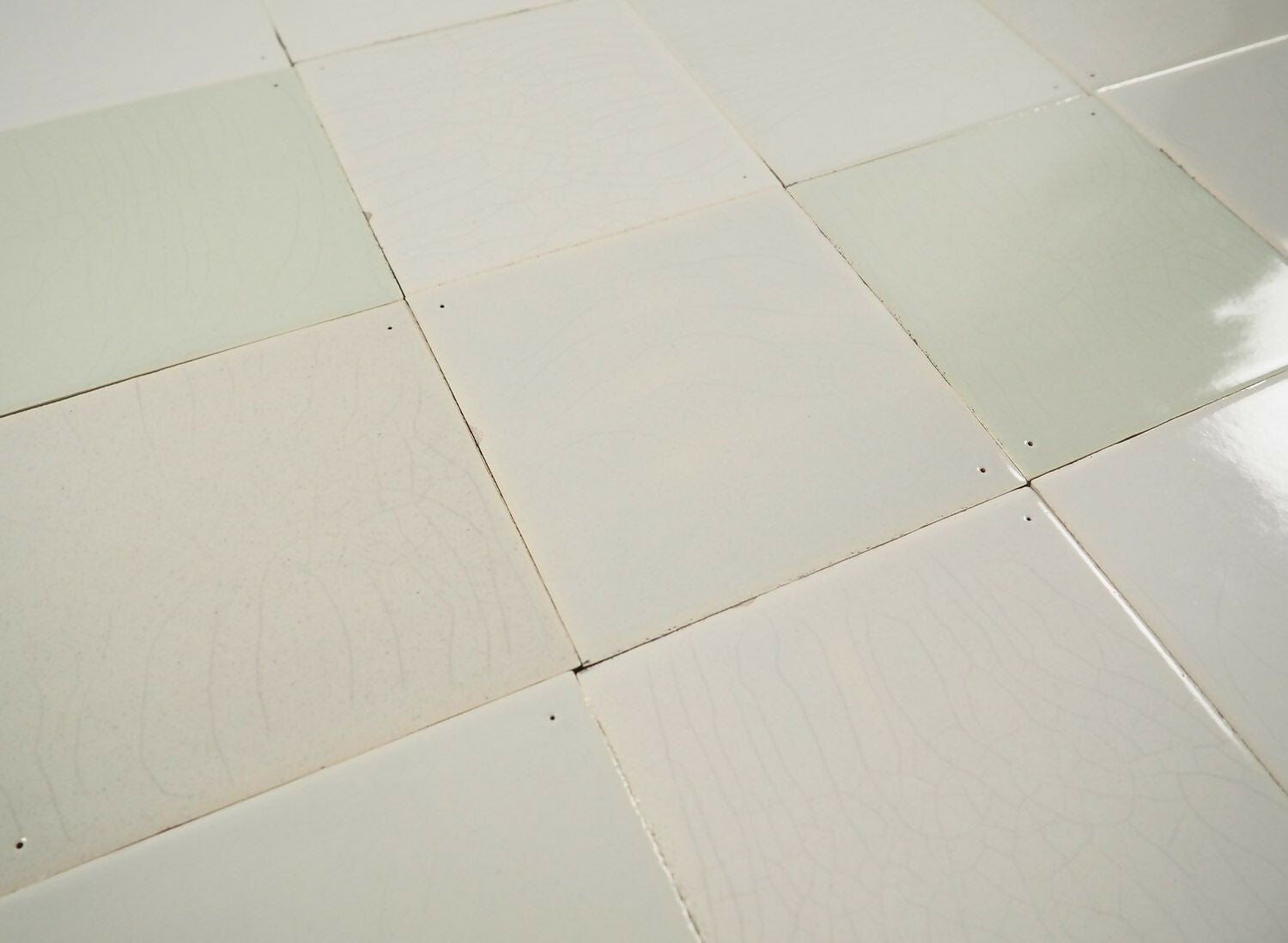 A grid of square Delft tiles in white, grey, and green shades, some with pinholes and a crackled finish, others without enhancements,  available  to buy on the-salon.com. SALON curates the finest , unique, handmade pieces from the interior industry's best makers and brands. Shop luxury furniture, lighting, rugs, art, antiques and vintage pieces and accessories from SALON interior designed rooms.