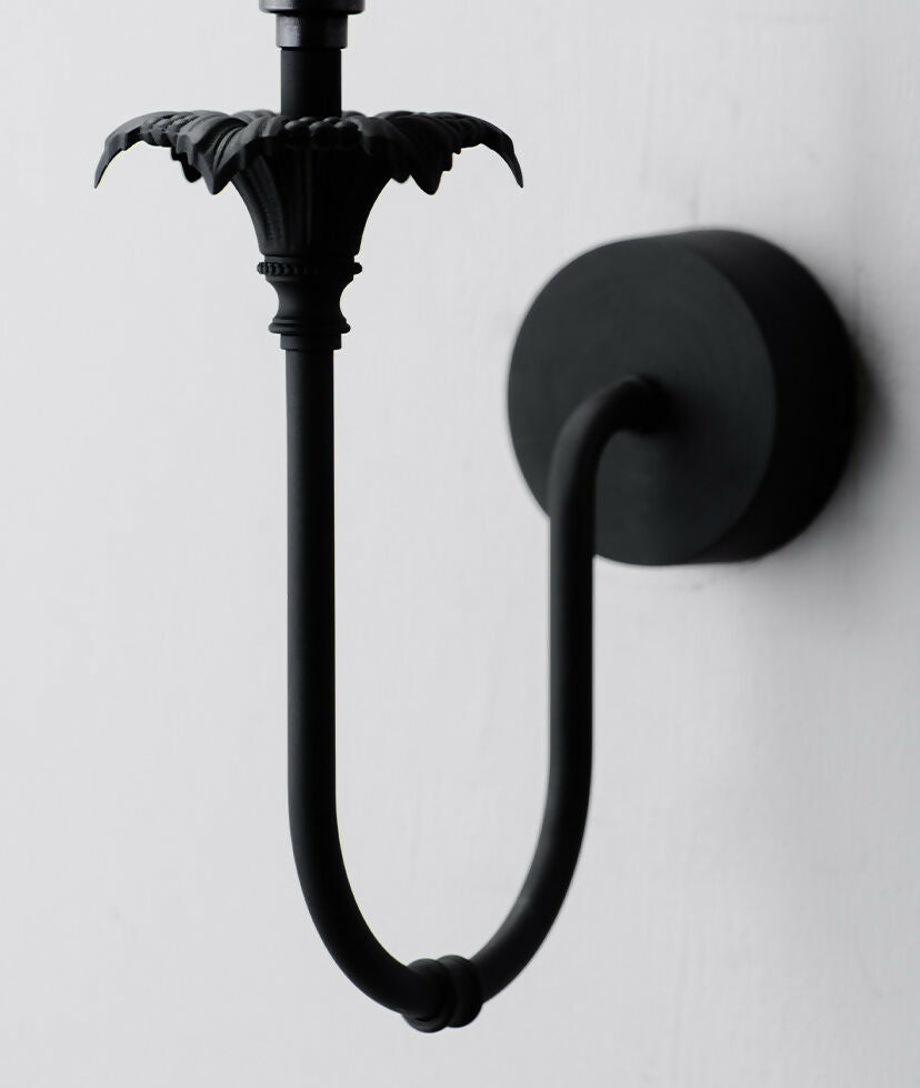 A wall-mounted light fixture with a black-colored metal arm and a white, solid-patterned lampshade,  available  to buy on the-salon.com. SALON curates the finest , unique, handmade pieces from the interior industry's best makers and brands. Shop luxury furniture, lighting, rugs, art, antiques and vintage pieces and accessories from SALON interior designed rooms.