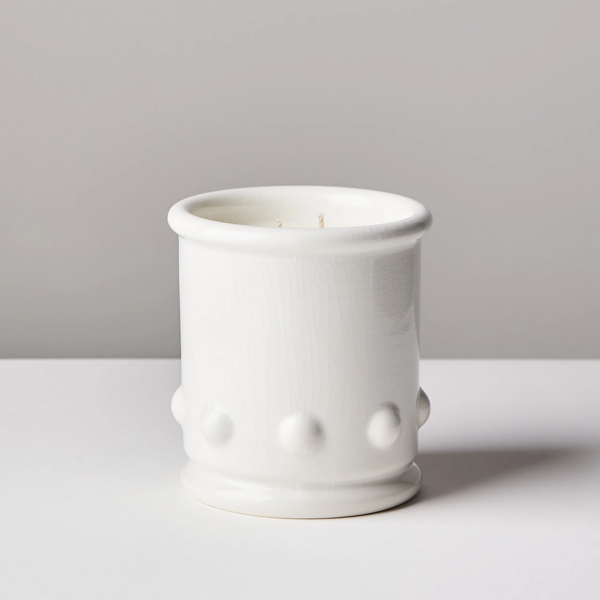 A white ceramic candle vessel with spherical detailing on the sides, with a candle visible at the top. The vessel is placed against a neutral background,  available  to buy on the-salon.com. SALON curates the finest , unique, handmade pieces from the interior industry's best makers and brands. Shop luxury furniture, lighting, rugs, art, antiques and vintage pieces and accessories from SALON interior designed rooms.