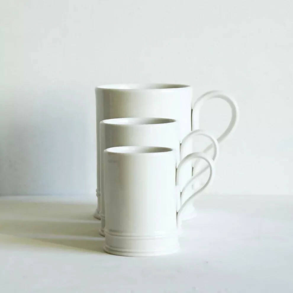 Three white porcelain coffee mugs, in a line with the biggest at the back and the smallest in the front, on a plain background, available  to buy on the-salon.com. SALON curates the finest , unique, handmade pieces from the interior industry's best makers and brands. Shop luxury furniture, lighting, rugs, art, antiques and vintage pieces and accessories from SALON interior designed rooms.