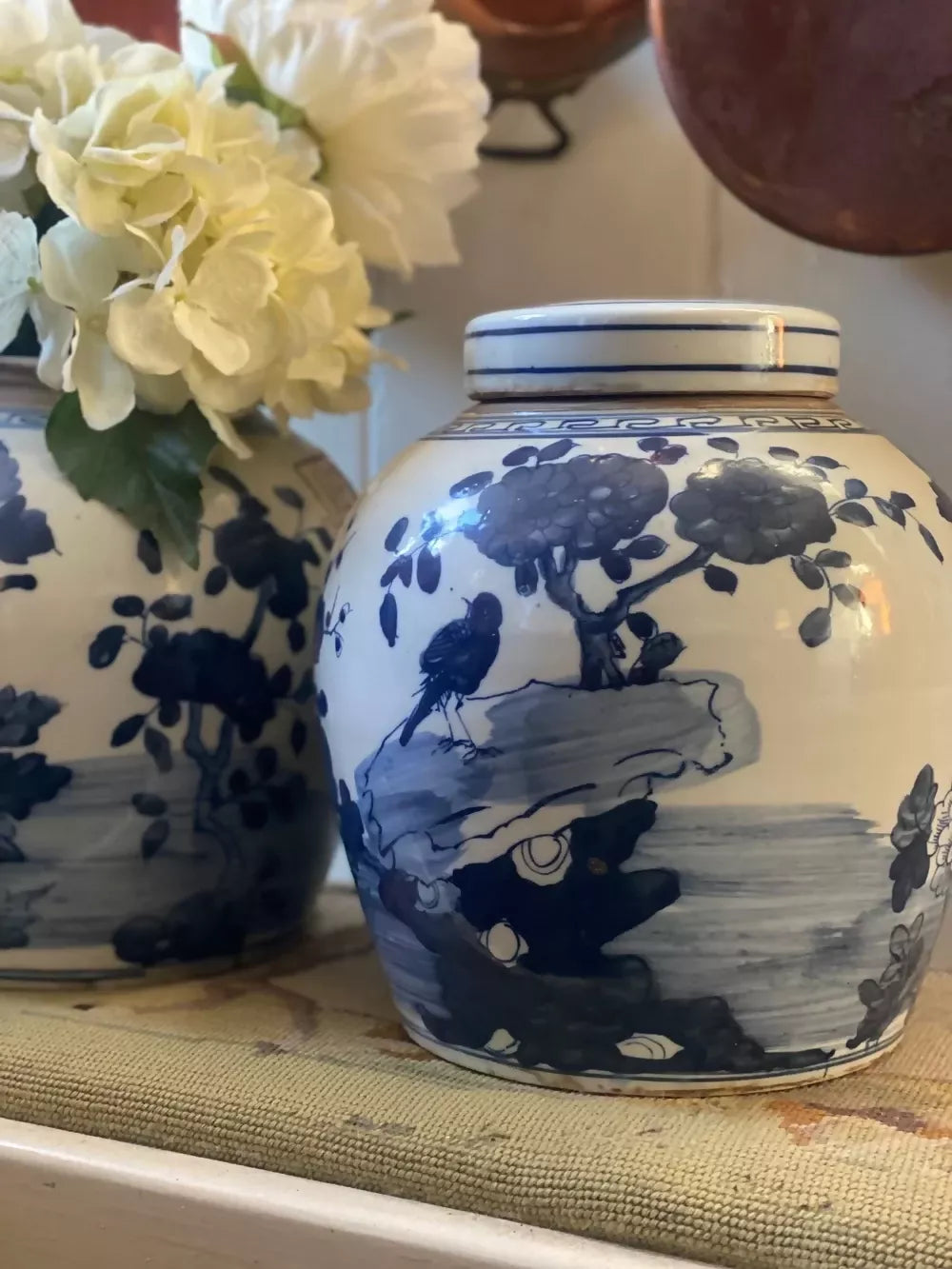 Blue and white ceramic jar with nature-themed design on a surface with flowers in the background,  available  to buy on the-salon.com. SALON curates the finest , unique, handmade pieces from the interior industry's best makers and brands. Shop luxury furniture, lighting, rugs, art, antiques and vintage pieces and accessories from SALON interior designed rooms.