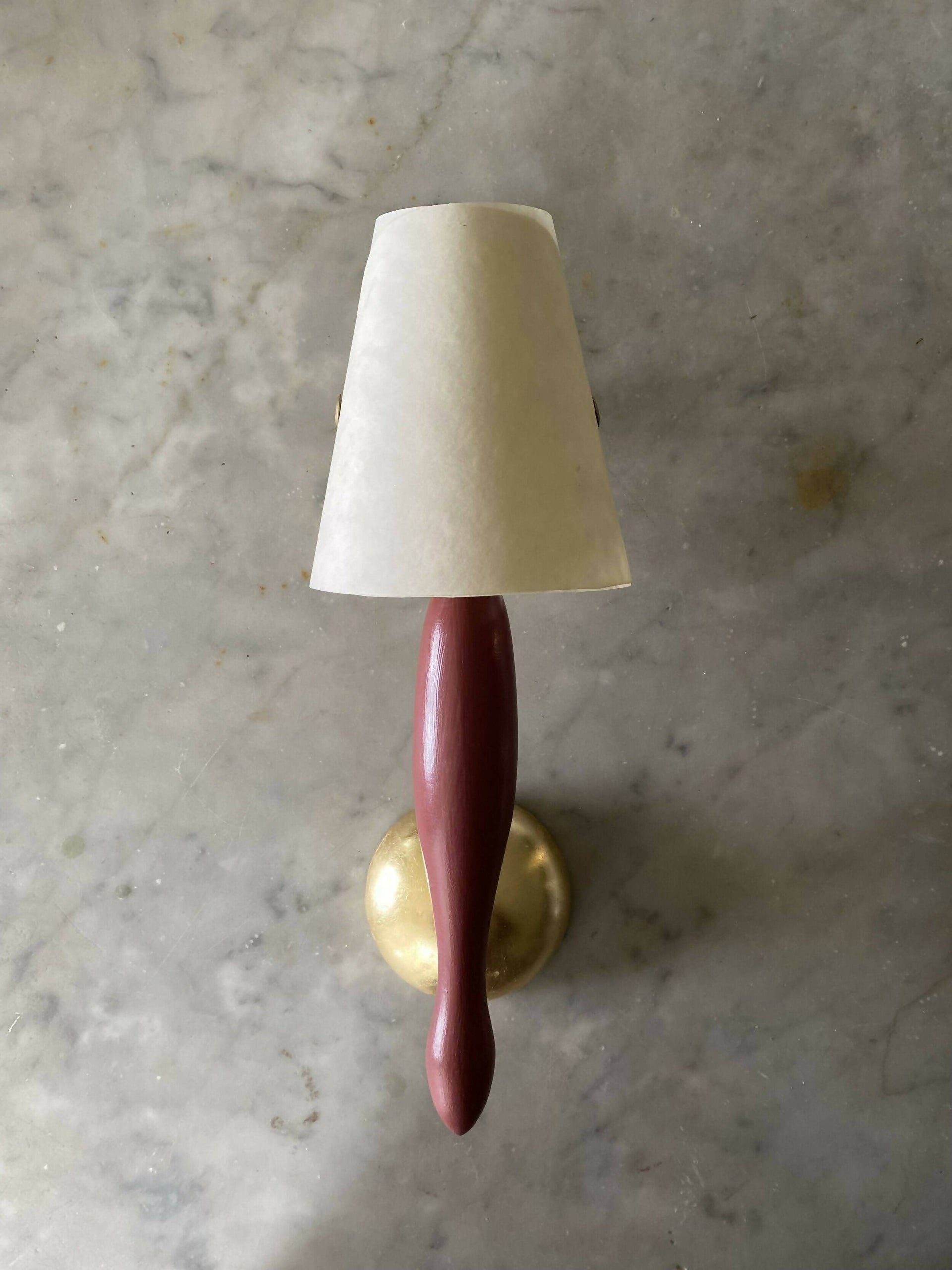 A wall light fixture with a burgundy, stem-like structure and a white conical shade, mounted on a gold-colored plate, available  to buy on the-salon.com. SALON curates the finest , unique, handmade pieces from the interior industry's best makers and brands. Shop luxury furniture, lighting, rugs, art, antiques and vintage pieces and accessories from SALON interior designed rooms.