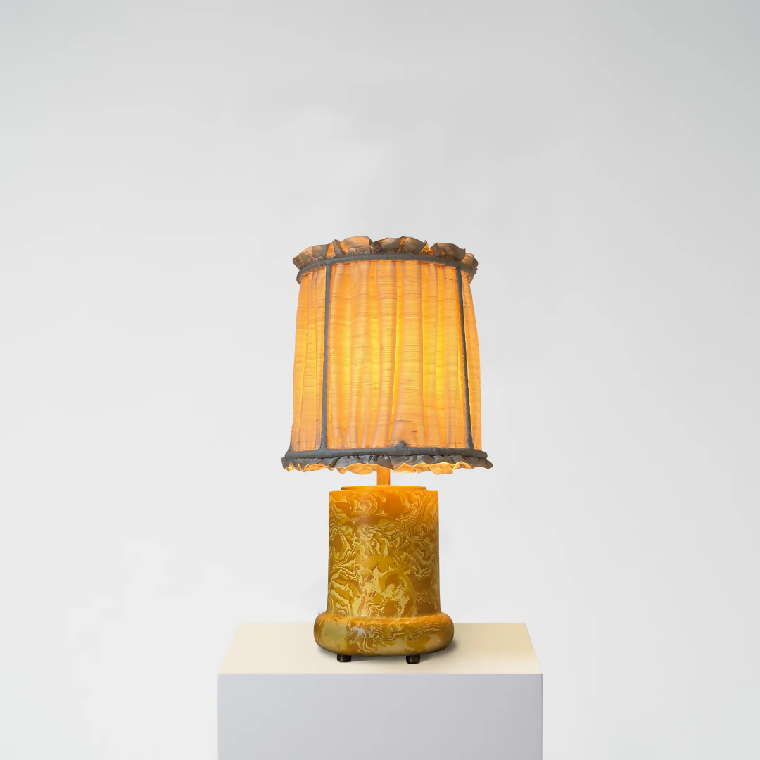 A yellow scagliola table lamp with a floral pattern base and a beige lampshade, displayed on a white platform against a neutral background, available  to buy on the-salon.com. SALON curates the finest , unique, handmade pieces from the interior industry's best makers and brands. Shop luxury furniture, lighting, rugs, art, antiques and vintage pieces and accessories from SALON interior designed rooms.