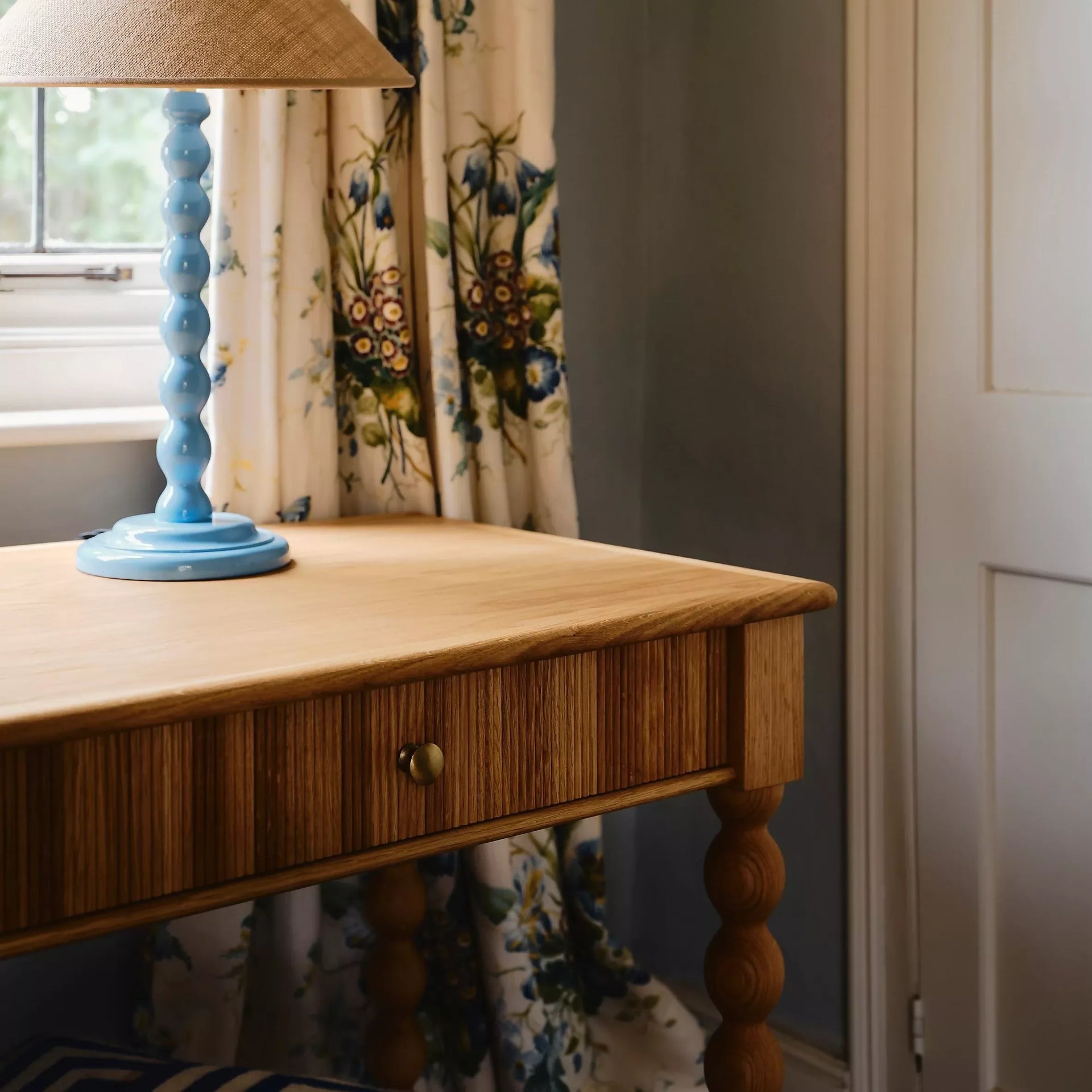 Wooden desk with a blue lamp on it, floral curtains in the background,available to buy from the-salon.com. SALON curates the finest unique handmade pieces from the interior industry's best makers and brands. Shop luxury furniture, lighting, rugs, art, antiques and vintage pieces and accessories from SALON interior designed rooms. 