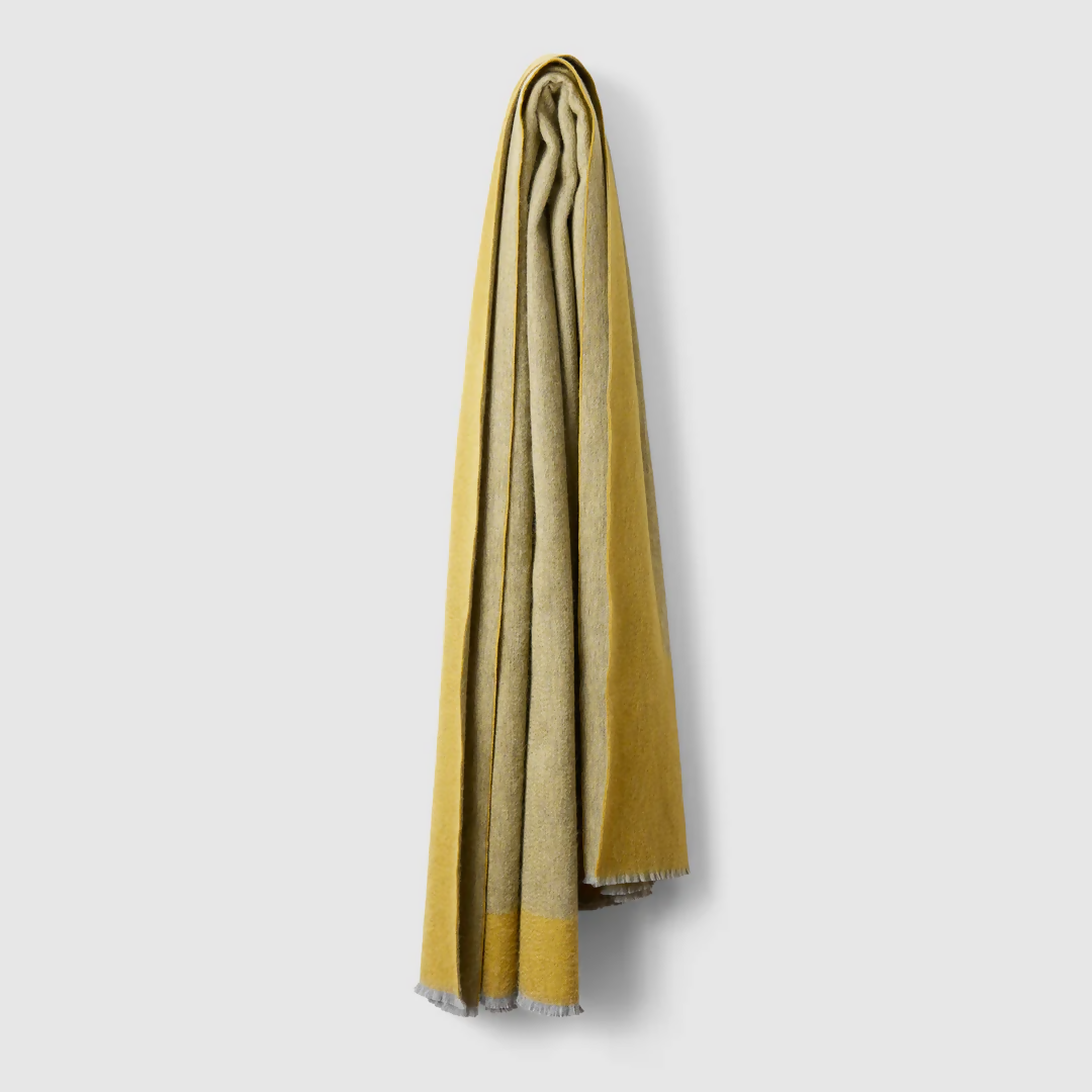 A gold-coloured cashmere throw blanket with a decorative border, displaying a small blue and silver brand tag on the bottom right corner,  available  to buy on the-salon.com. SALON curates the finest , unique, handmade pieces from the interior industry's best makers and brands. Shop luxury furniture, lighting, rugs, art, antiques and vintage pieces and accessories from SALON interior designed rooms.