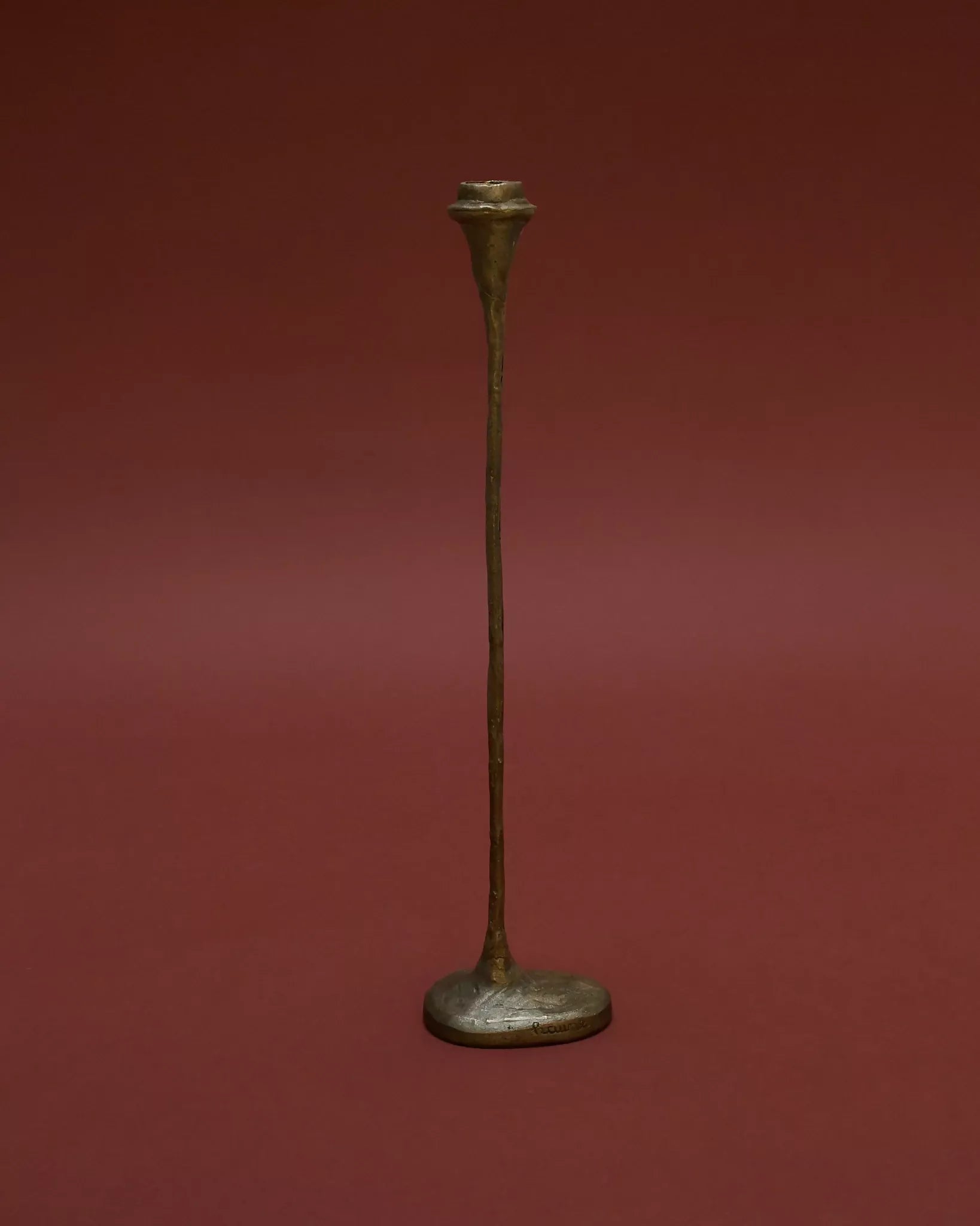 A tall, solid bronze candle holder with a textured finish, standing on a flat surface with a red background,  available  to buy on the-salon.com. SALON curates the finest , unique, handmade pieces from the interior industry's best makers and brands. Shop luxury furniture, lighting, rugs, art, antiques and vintage pieces and accessories from SALON interior designed rooms.