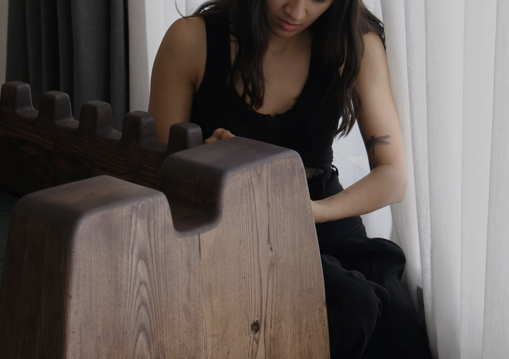 A woman sitting infront of  the wooden seat, highlighting the wooden stains on the chair,  available  to buy on the-salon.com. SALON curates the finest , unique, handmade pieces from the interior industry's best makers and brands. Shop luxury furniture, lighting, rugs, art, antiques and vintage pieces and accessories from SALON interior designed rooms.