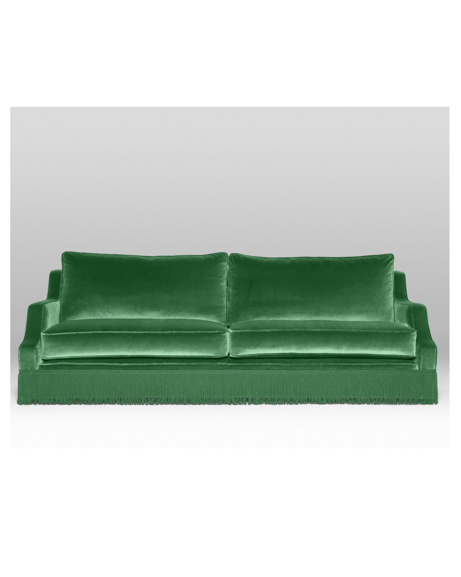 A green velvet sofa with plush seats and a tasseled fringe, named 'Large Atlanta Sofa by Orior', positioned in a room with a bookshelf, a chair, and a decorative rug,  available  to buy on the-salon.com. SALON curates the finest , unique, handmade pieces from the interior industry's best makers and brands. Shop luxury furniture, lighting, rugs, art, antiques and vintage pieces and accessories from SALON interior designed rooms.
