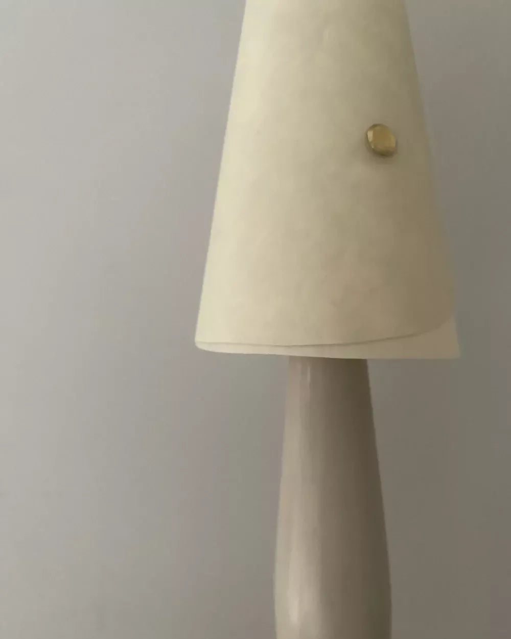 A close-up of a table lamp featuring a tapered off-white fabric shade with a small gold button detail and a matching off-white ceramic base, set against a plain light gray wall.