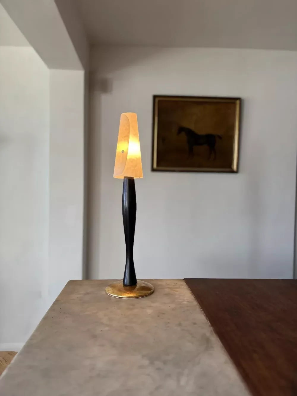 A modern table lamp with a black twisted base, a warm glowing tapered shade, and a gold circular base, placed on a stone countertop in a minimalist room.