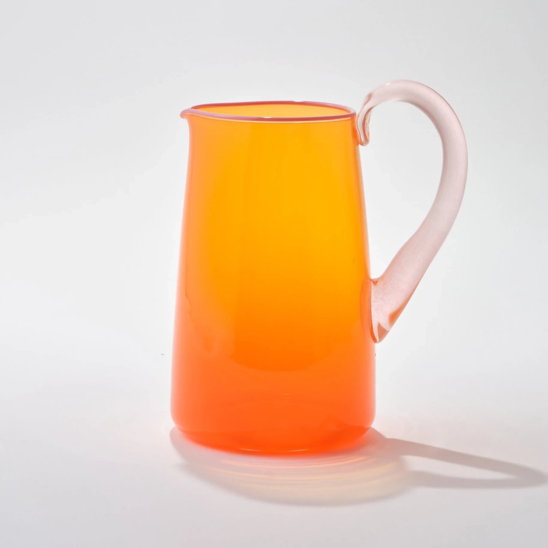 Hand blown, transparent orange glass jug with a blush handle and contrasting pale pink lip on a white background,  available  to buy on the-salon.com. SALON curates the finest , unique, handmade pieces from the interior industry's best makers and brands. Shop luxury furniture, lighting, rugs, art, antiques and vintage pieces and accessories from SALON interior designed rooms.