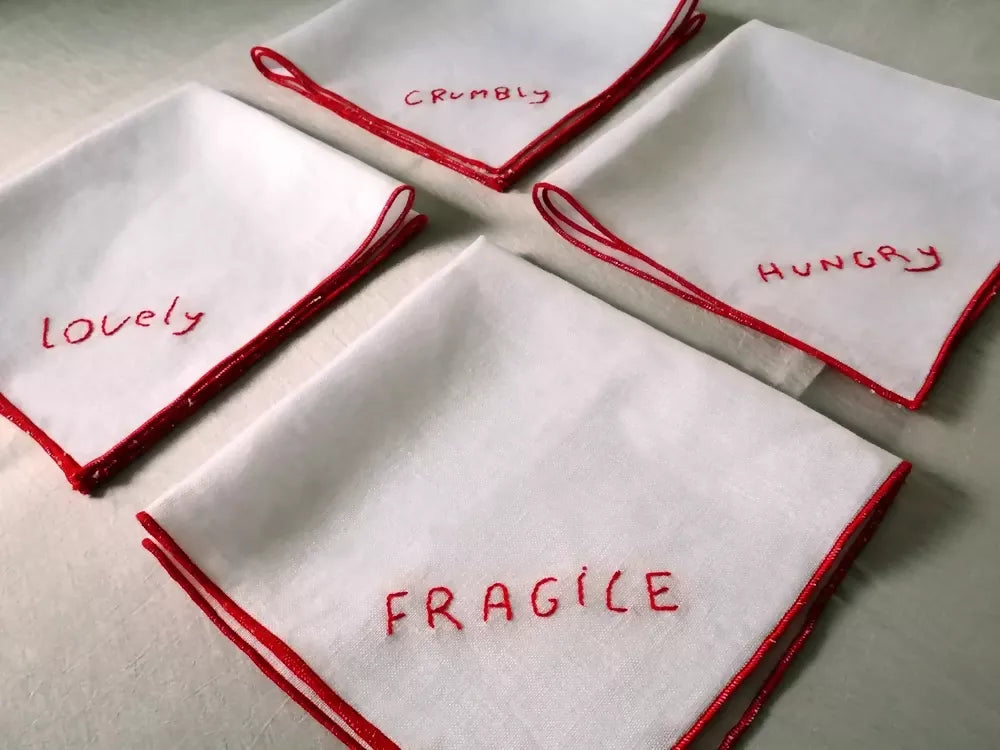 Four white napkins with red embroidered text on a light background,  available  to buy on the-salon.com. SALON curates the finest , unique, handmade pieces from the interior industry's best makers and brands. Shop luxury furniture, lighting, rugs, art, antiques and vintage pieces and accessories from SALON interior designed rooms.