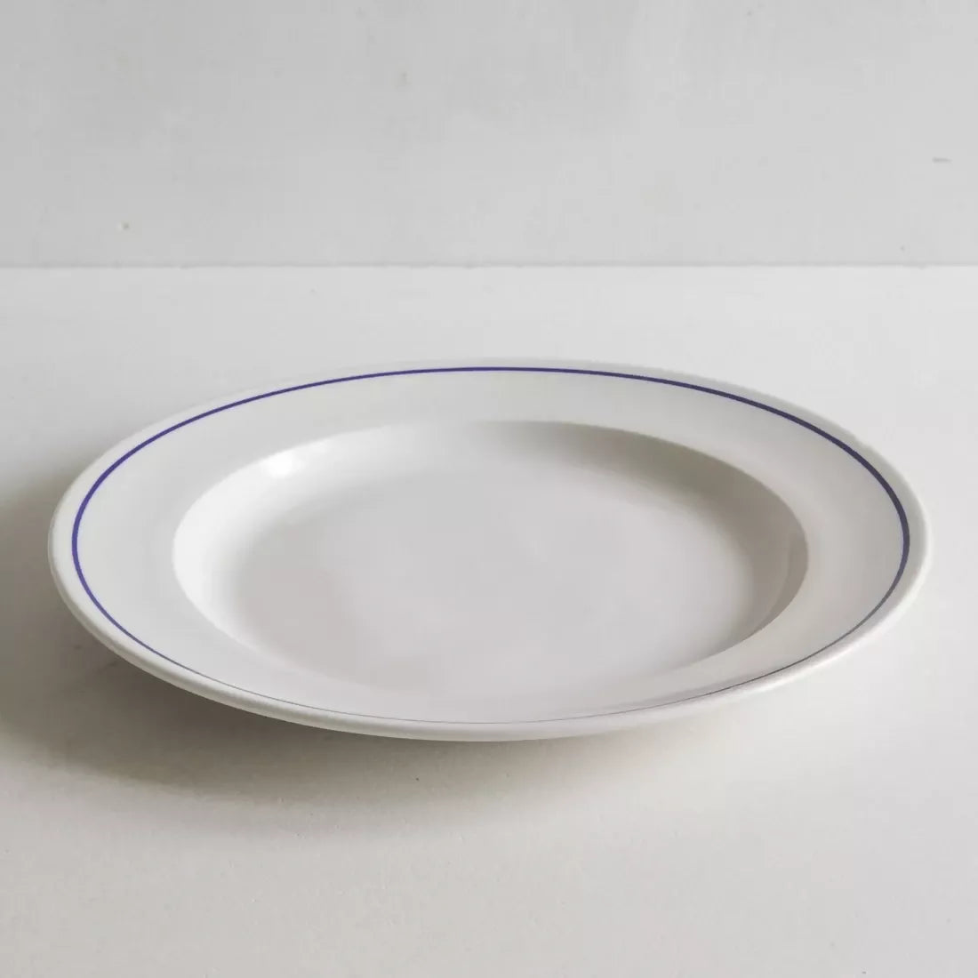 A white porcelain dinner plate with a rich cobalt blue line along the rim, placed against a neutral background, available  to buy on the-salon.com. SALON curates the finest , unique, handmade pieces from the interior industry's best makers and brands. Shop luxury furniture, lighting, rugs, art, antiques and vintage pieces and accessories from SALON interior designed rooms.