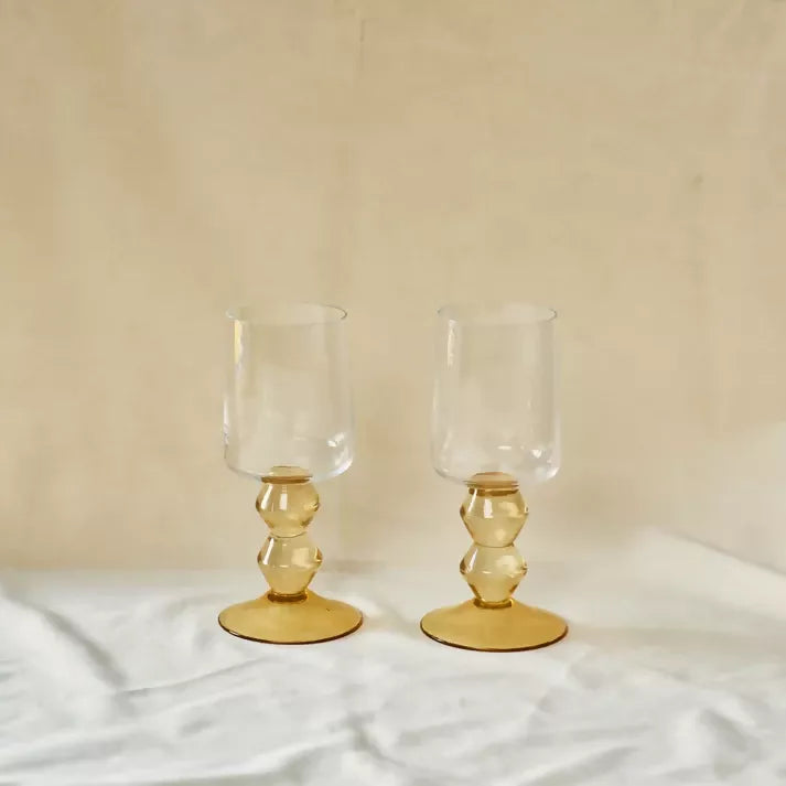 Two clear glasses with gold bases on a light fabric background, available  to buy on the-salon.com. SALON curates the finest , unique, handmade pieces from the interior industry's best makers and brands. Shop luxury furniture, lighting, rugs, art, antiques and vintage pieces and accessories from SALON interior designed rooms.