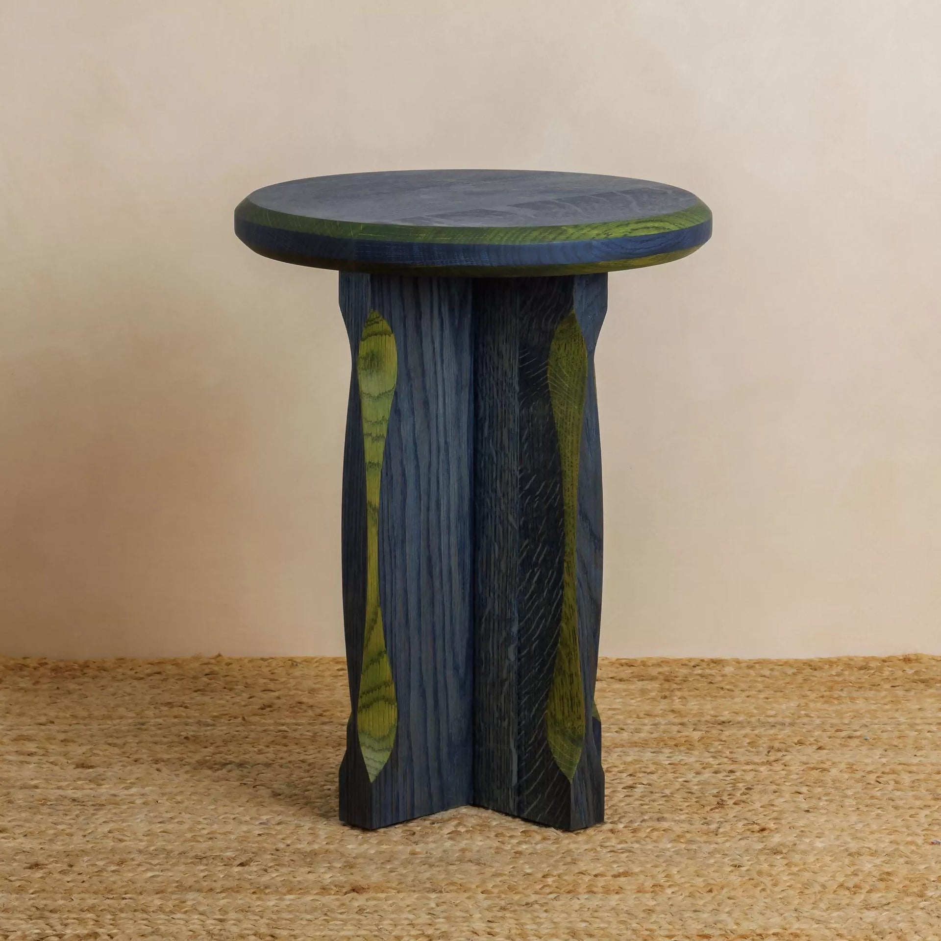 A round, blue with green oak side table with a cross base, placed on a woven mat,  available  to buy on the-salon.com. SALON curates the finest , unique, handmade pieces from the interior industry's best makers and brands. Shop luxury furniture, lighting, rugs, art, antiques and vintage pieces and accessories from SALON interior designed rooms.
