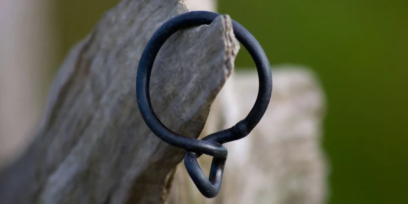 A black wrought iron curtain ring hanging on a wooden surface, with a natural outdoor background,  available  to buy on the-salon.com. SALON curates the finest , unique, handmade pieces from the interior industry's best makers and brands. Shop luxury furniture, lighting, rugs, art, antiques and vintage pieces and accessories from SALON interior designed rooms.